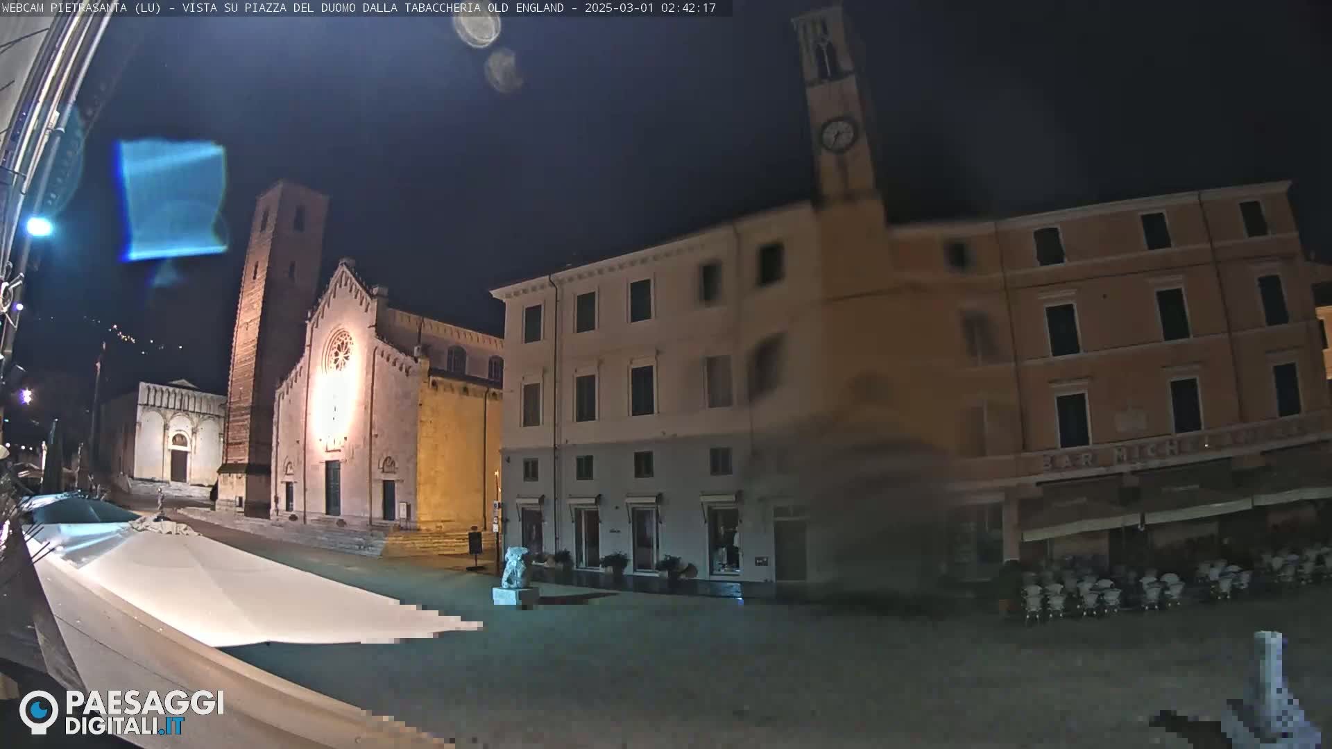 A nighttime view of a town square, featuring a church, buildings, and a statue, under a dark, possibly cloudy sky.
