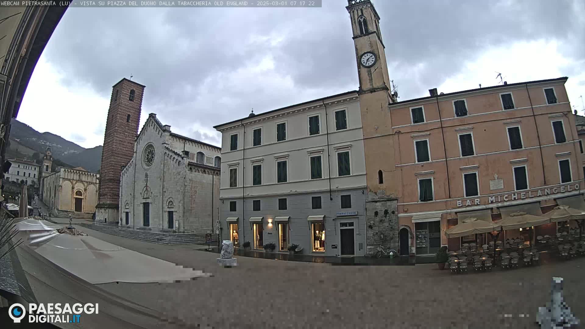 A town square with stone buildings, a church, and a clock tower under an overcast sky.