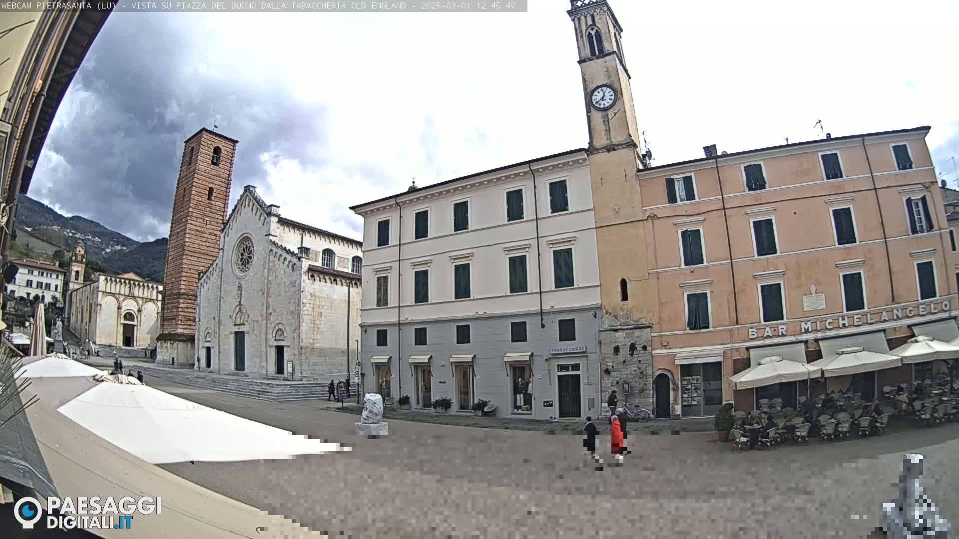 A town square is shown under a cloudy sky, featuring a large church with a tall bell tower, multi-story buildings with awnings, and a few people walking.