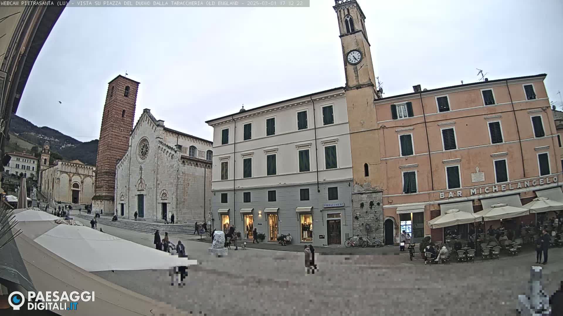 A town square, under an overcast sky, features a large stone church, several buildings, outdoor seating areas, and a few people.