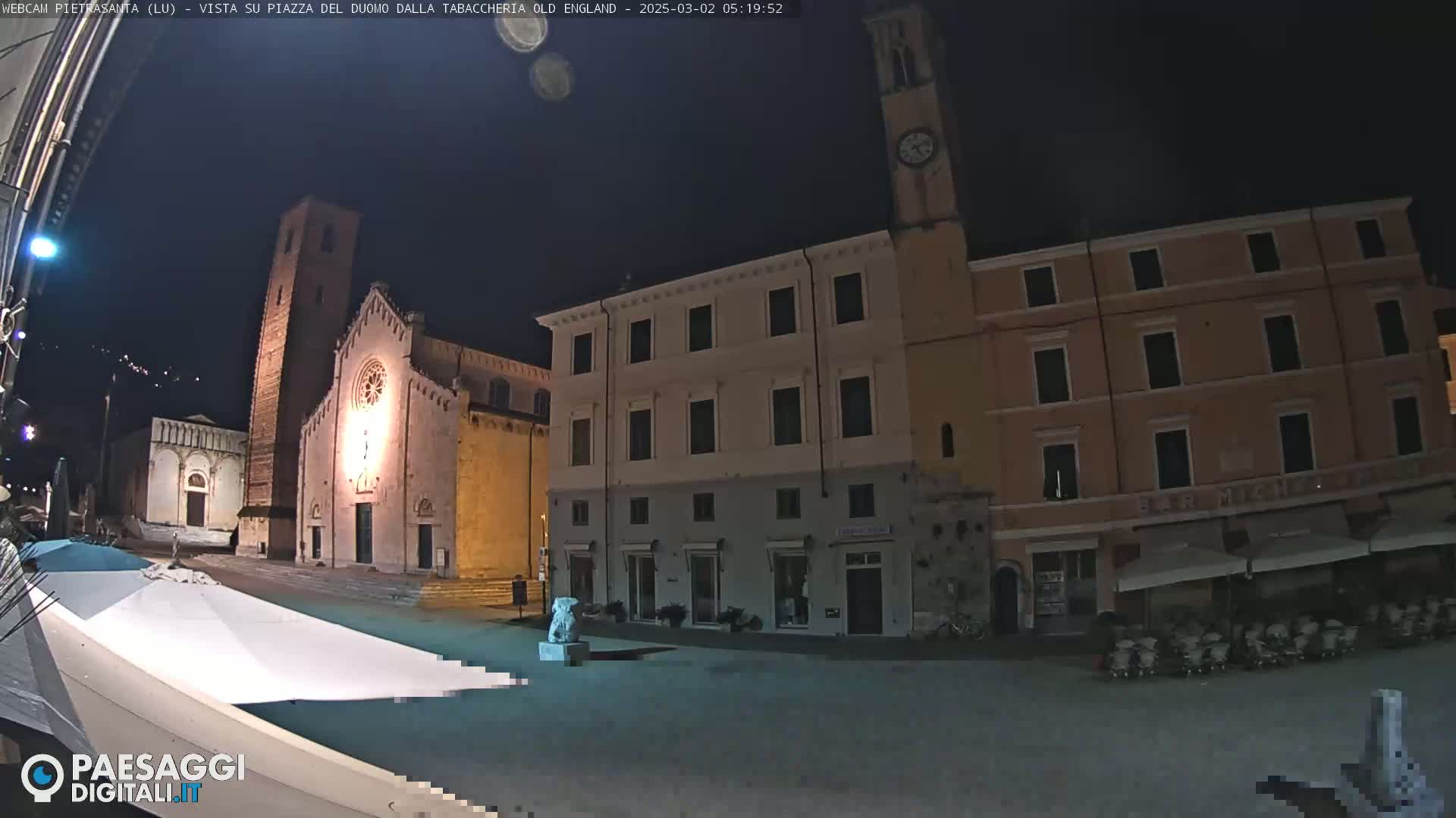 A nighttime view of a town square with buildings, a church, and a clock tower under a dark, clear sky.