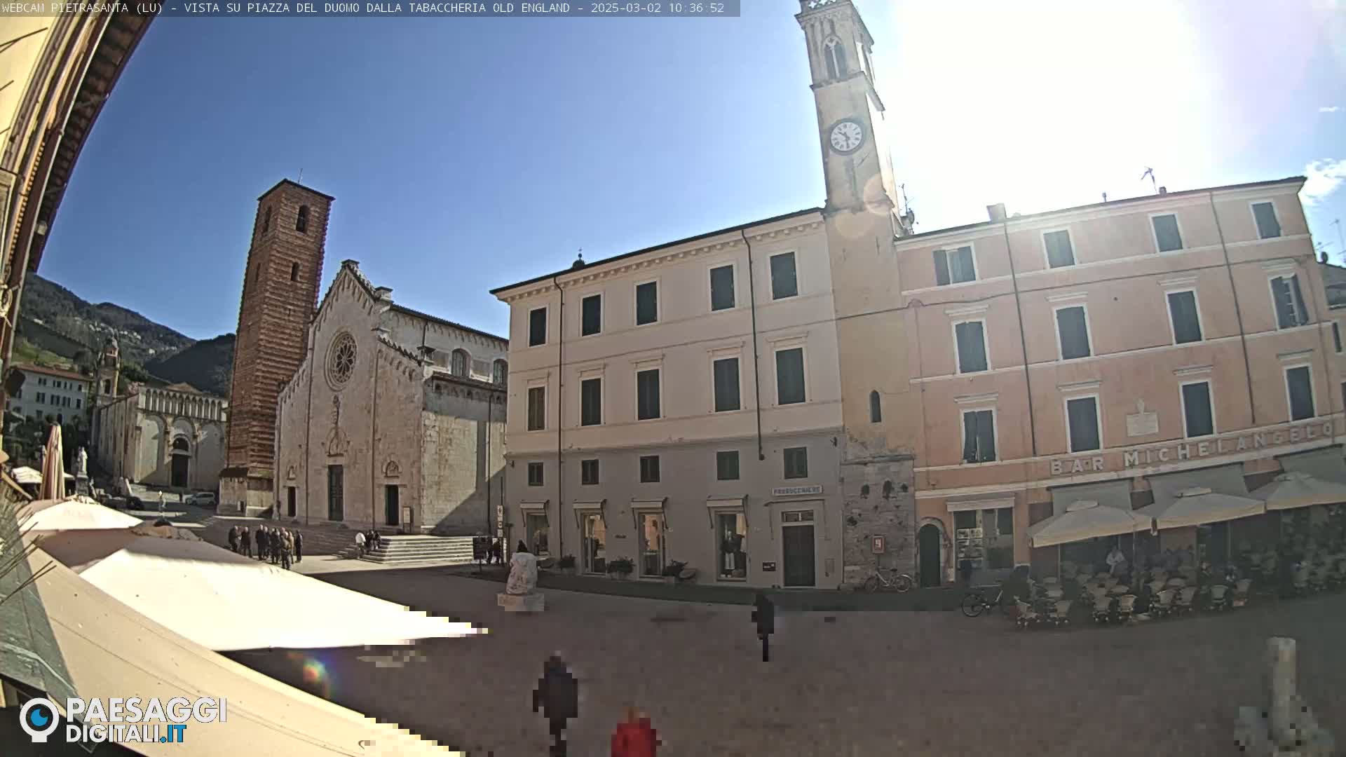 A sunny day in a town square shows a large cathedral and other buildings, with people walking around and some outdoor seating under umbrellas.