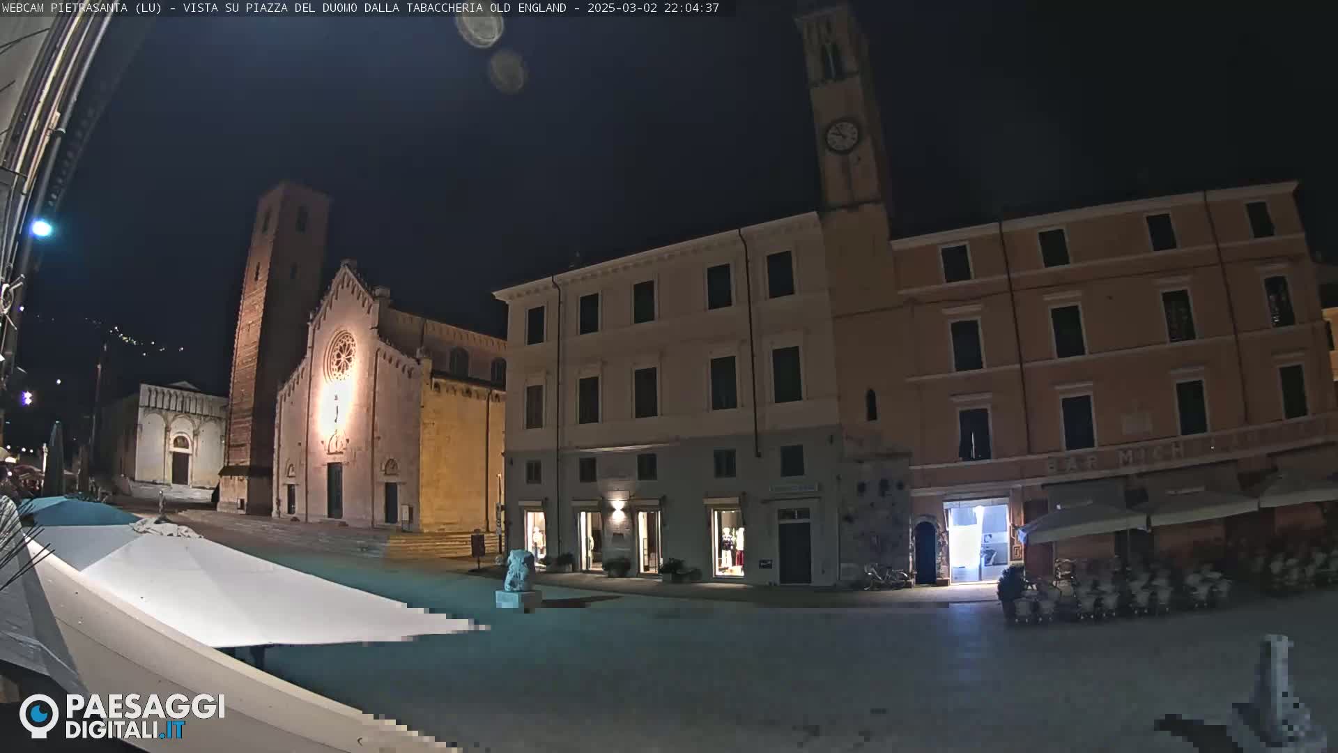 A nighttime view of a town square with a church, several buildings, and outdoor seating under covered areas.