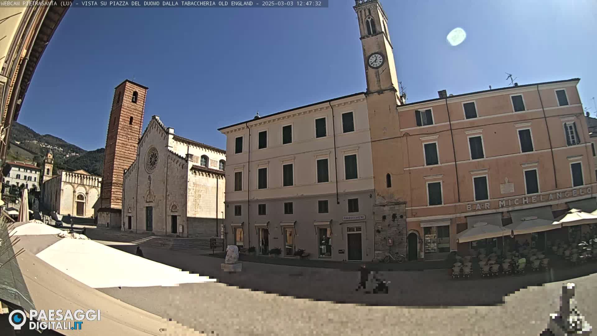 A sunny town square features a large church with a tall bell tower, adjacent buildings, and outdoor seating at a café.