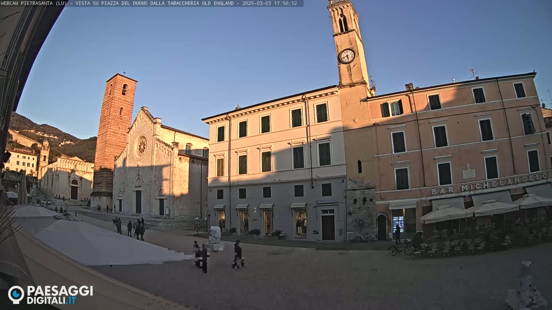 A sunlit town square is seen with several buildings, including a church and a clock tower, under a clear blue sky.