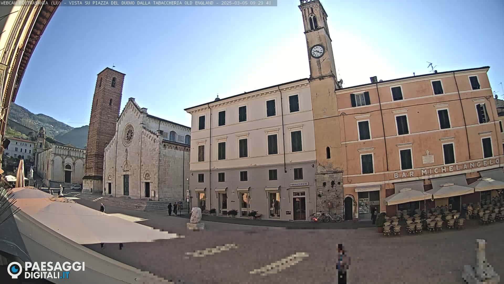 A sunny view of a town square with a large cathedral, a clock tower, and several buildings, including a cafe with outdoor seating.