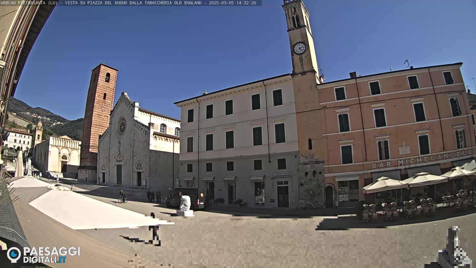 A sunny town square is shown, featuring a large cathedral, several buildings, a clock tower, and outdoor seating at a café.