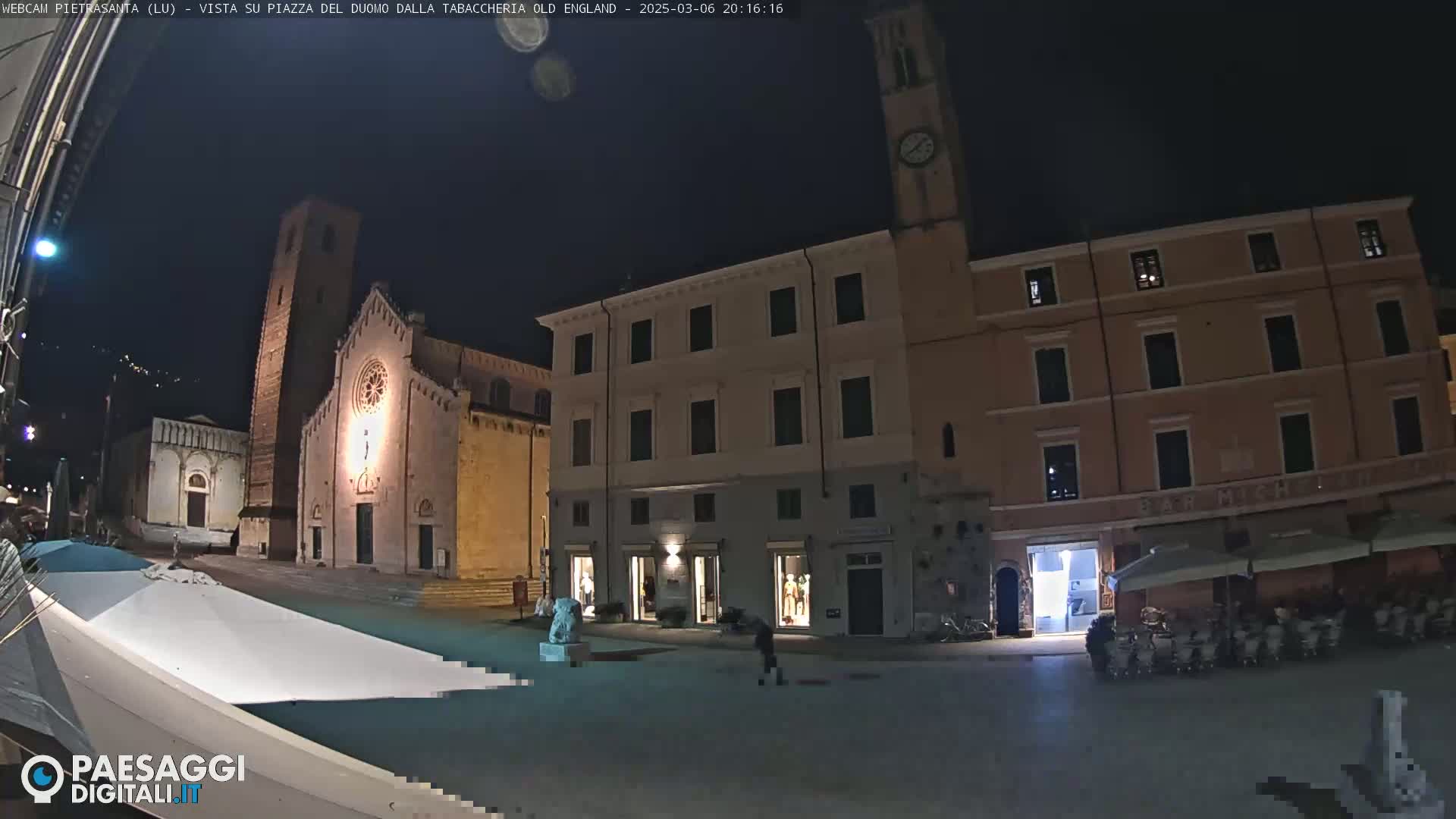 A nighttime view of a town square with a church, buildings, and a few people, under a clear, dark sky.