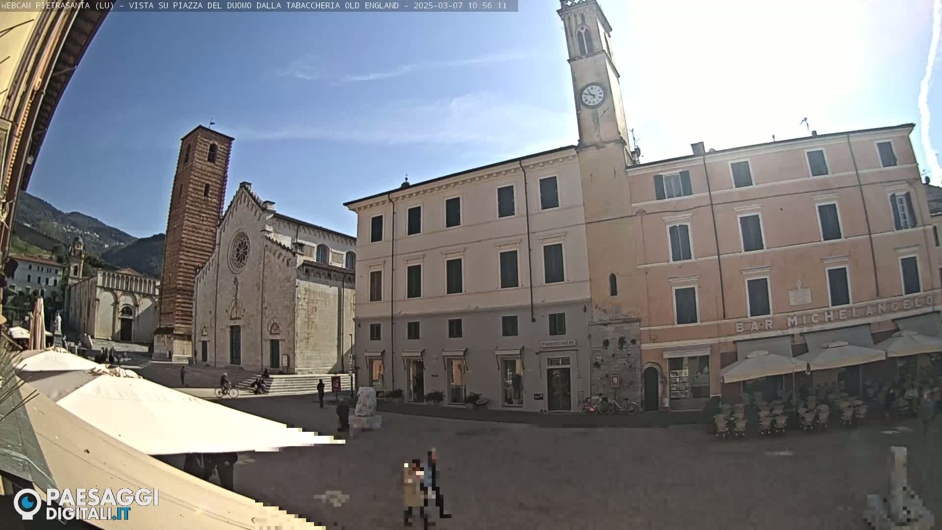 A sunny town square with a large cathedral and several multi-story buildings, including a bar, is shown in the image.