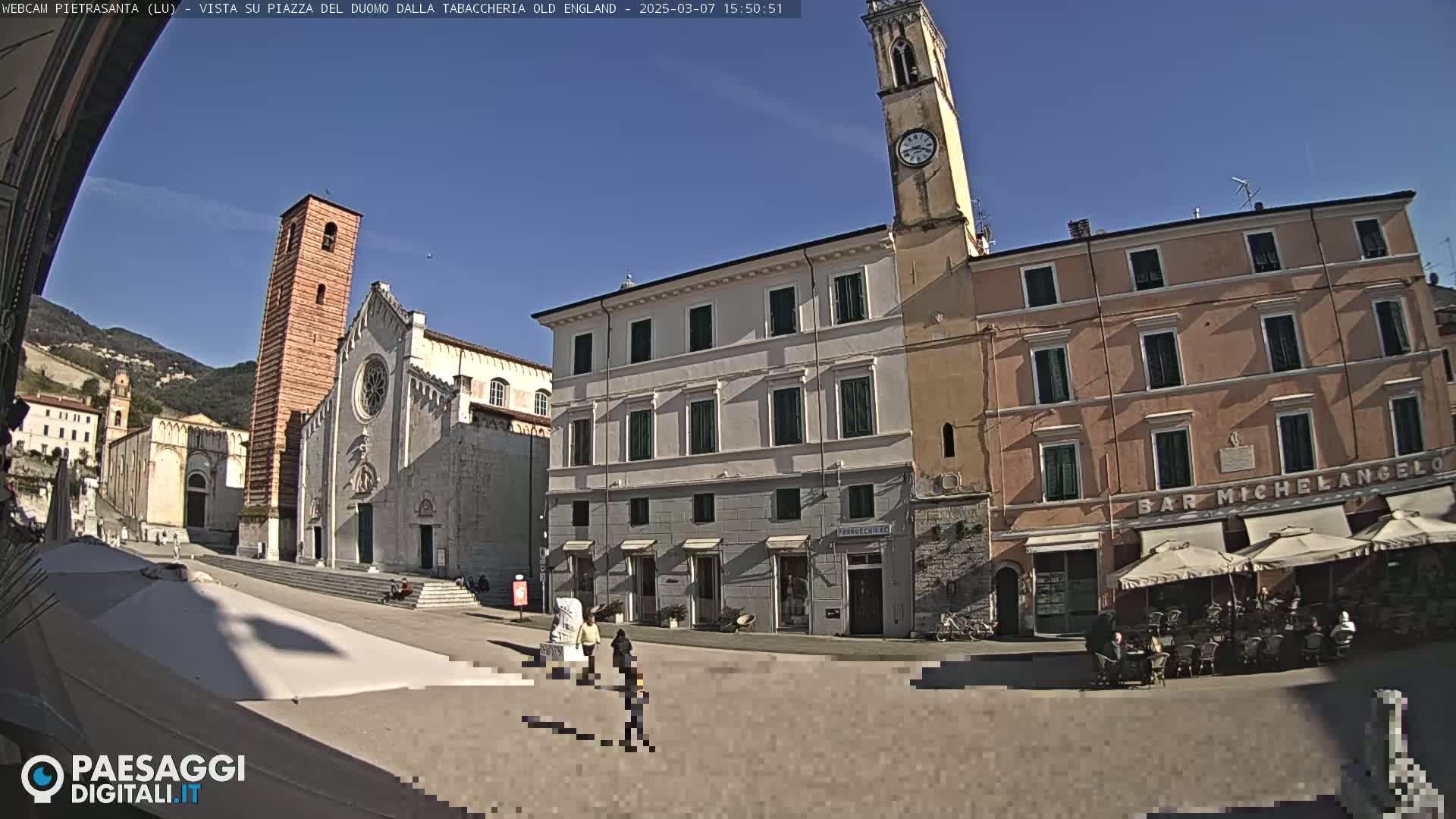 A sunny town square with a large church, clock tower, and several buildings, including a café with outdoor seating.