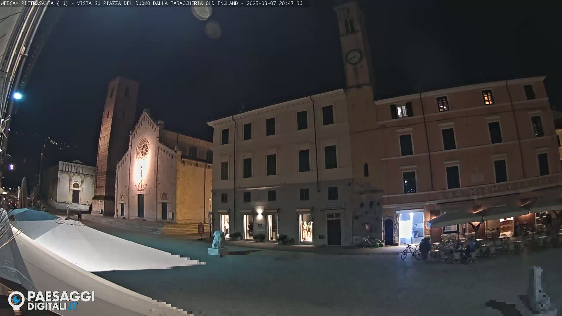 A town square at night, illuminated by artificial light, features a church with a tall bell tower, several buildings, and a few people sitting at outdoor tables.
