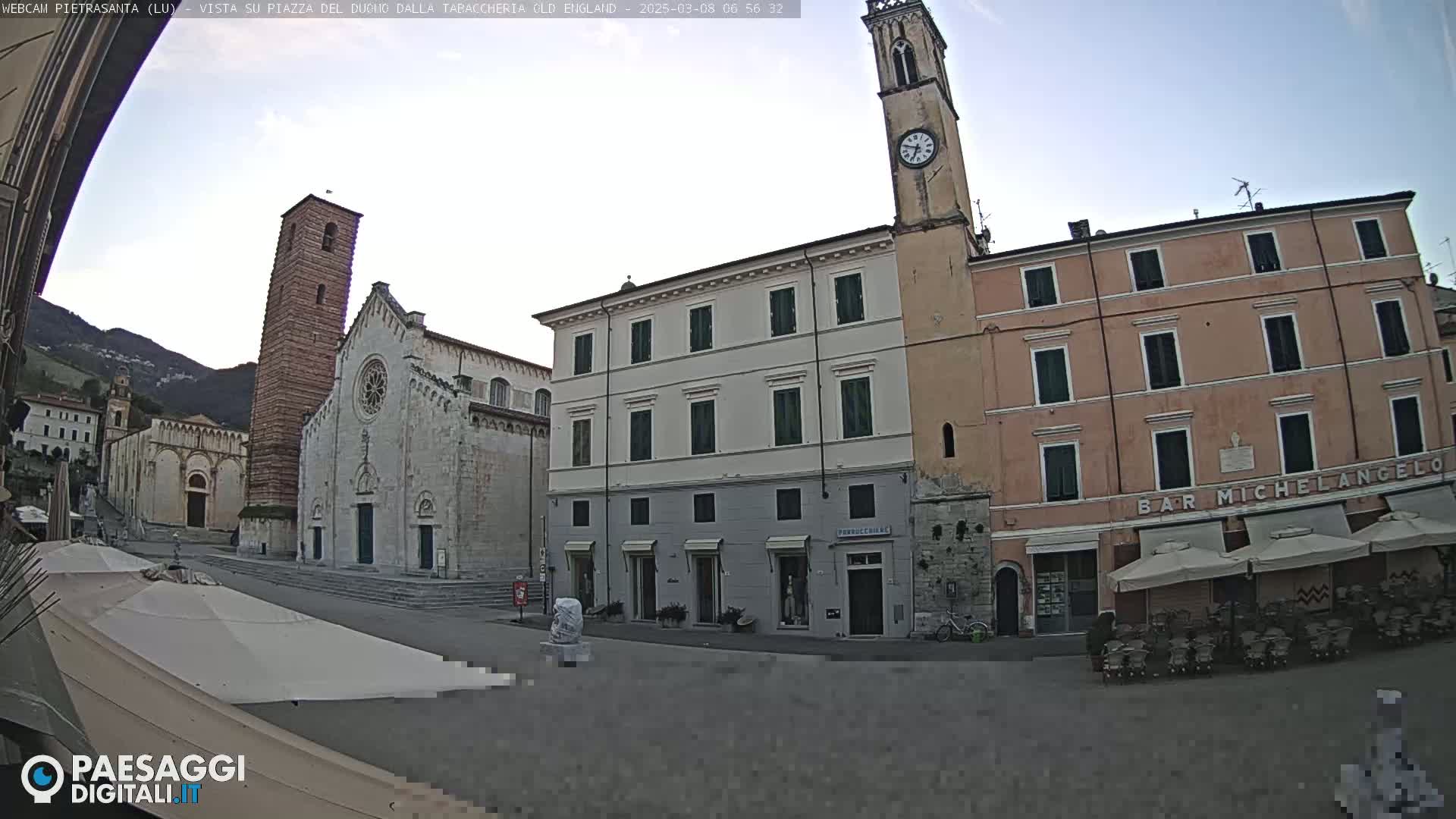 A mostly clear day shows an Italian town square with a large church, several buildings, and outdoor seating areas under umbrellas.
