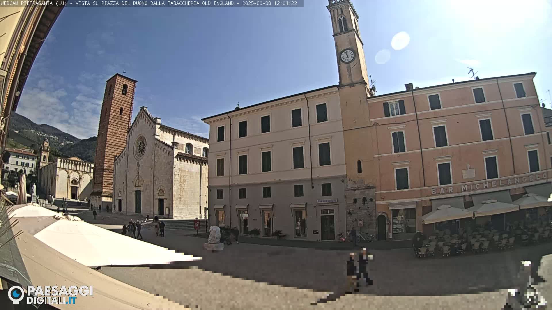 A sunny town square features a large cathedral, several multi-story buildings, and outdoor seating under umbrellas.