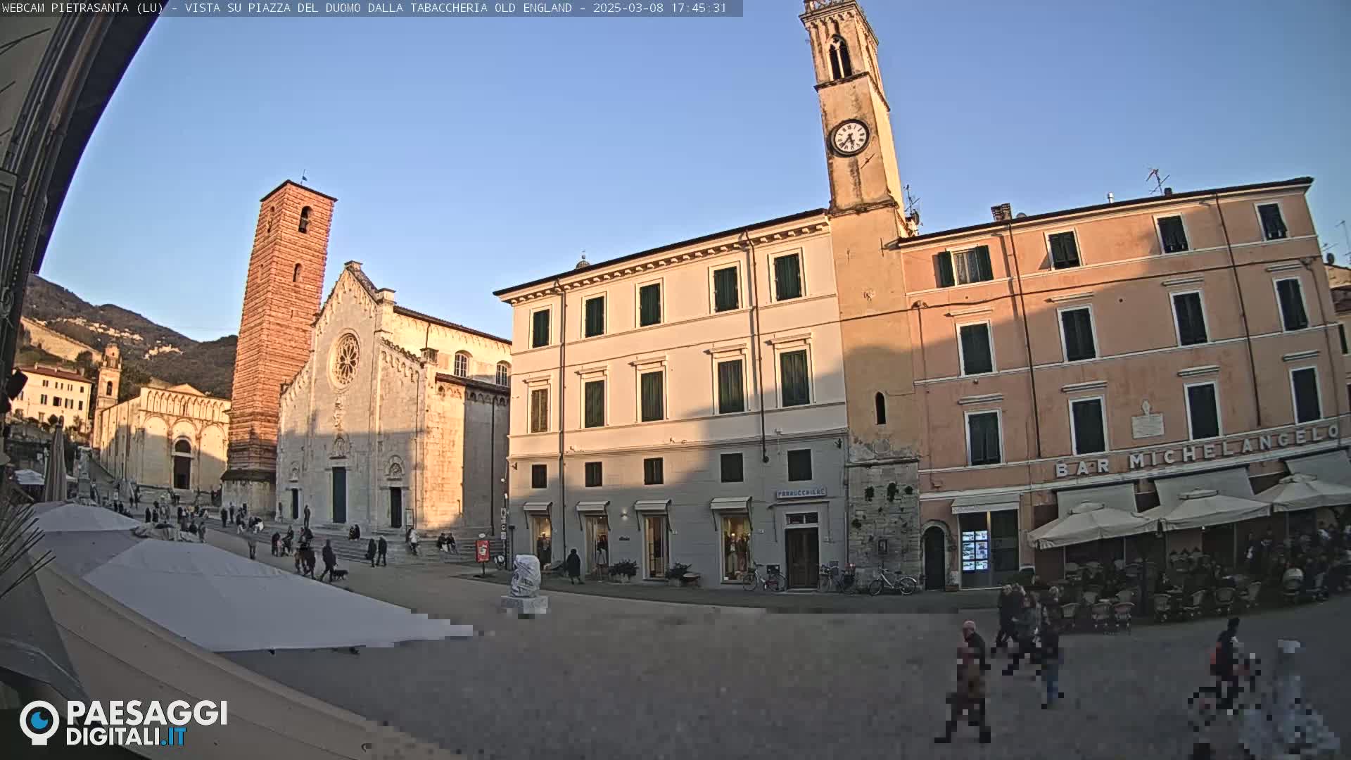 A town square in the late afternoon, under a clear blue sky, is surrounded by historical buildings, including a cathedral and a clock tower, with people walking around and several outdoor cafe umbrellas set up.