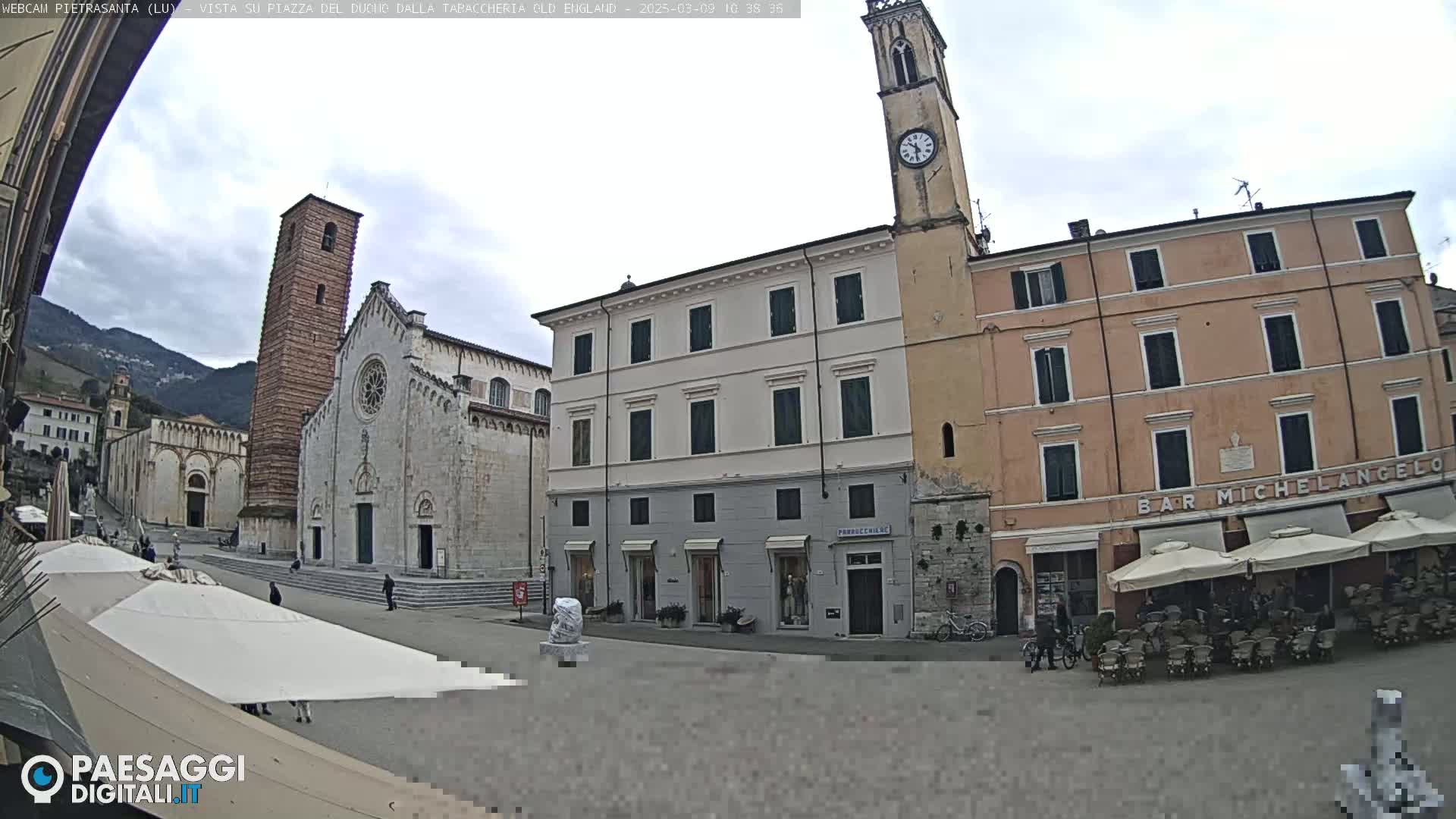 A town square, under an overcast sky, features a large stone cathedral, multi-story buildings, and outdoor seating areas with umbrellas.