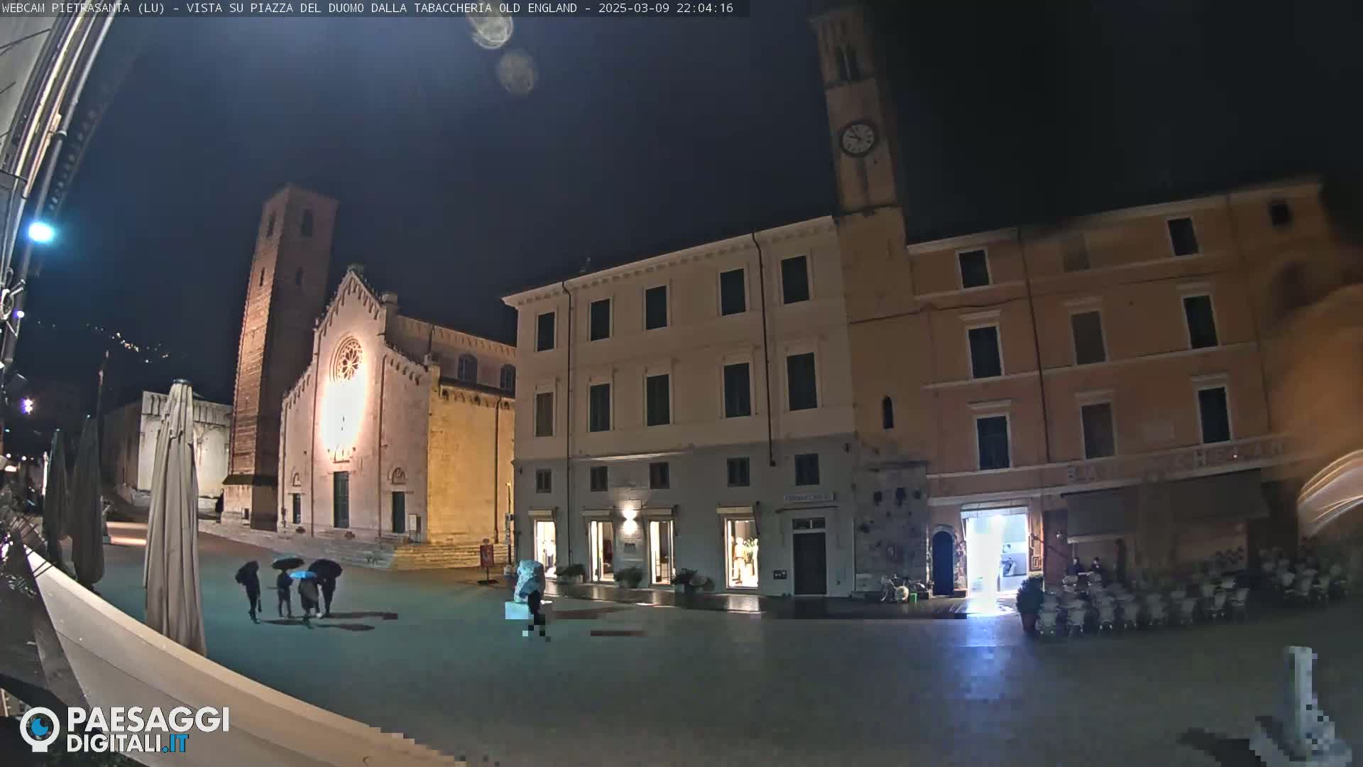A nighttime view of a town square with several buildings, including a church, under a clear, dark sky, with a few people walking around.