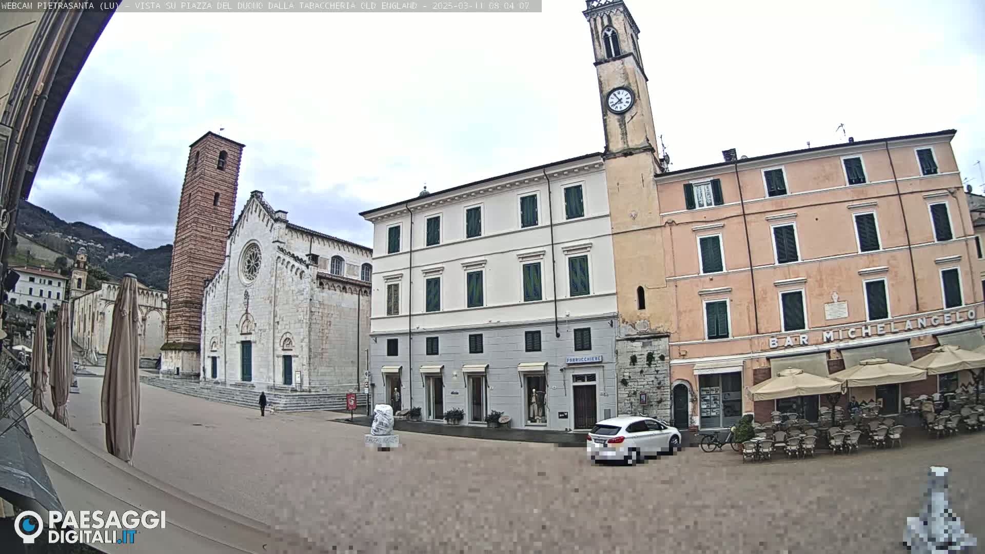 A town square with a church, several buildings, a cafe with outdoor seating, and a car, all under an overcast sky.
