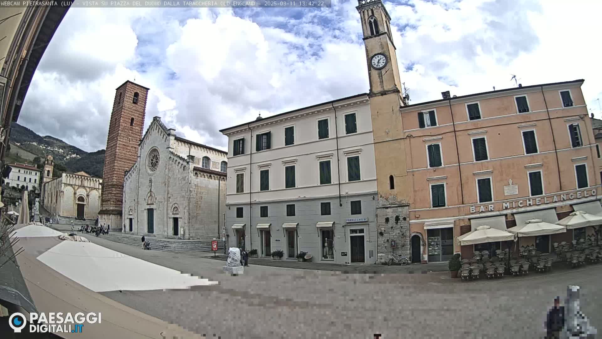 A town square with a large church, several buildings, and outdoor seating under umbrellas, under partly cloudy skies.