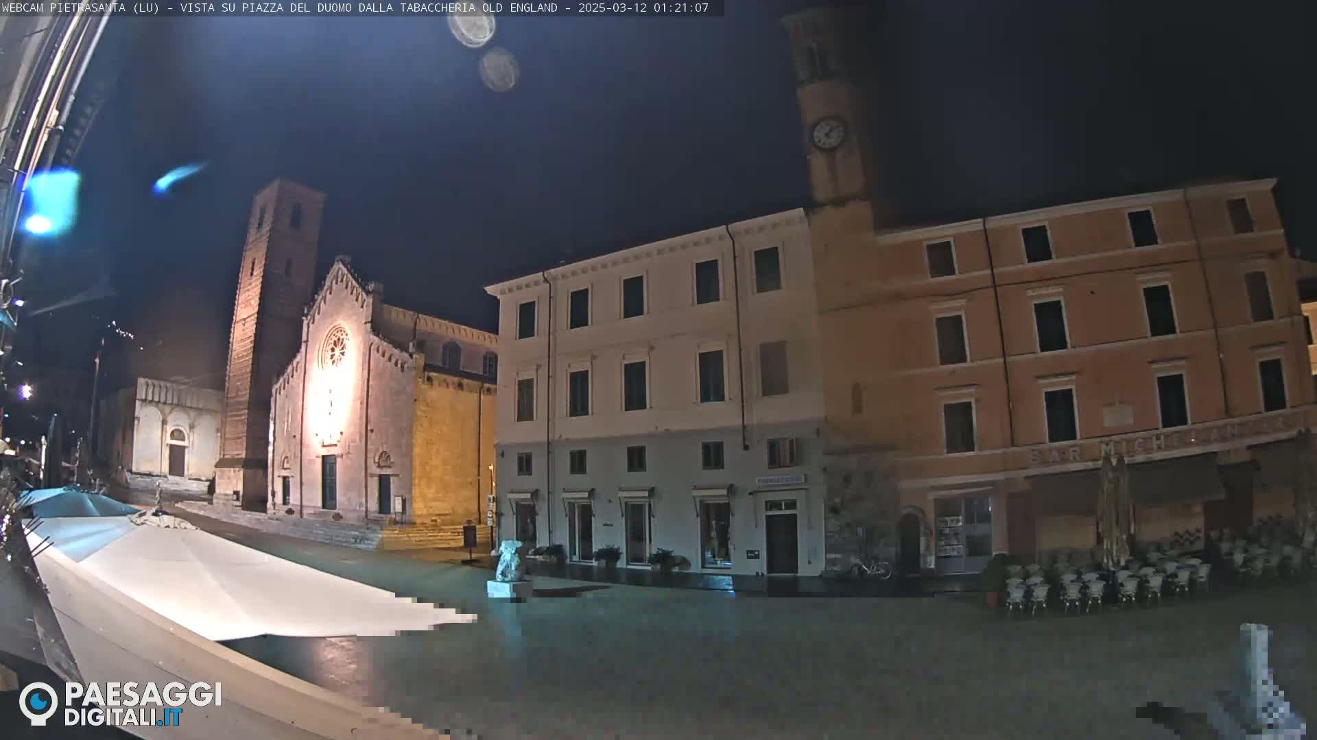 A nighttime view of a town square with a church, buildings, and outdoor seating under a dark, clear sky.