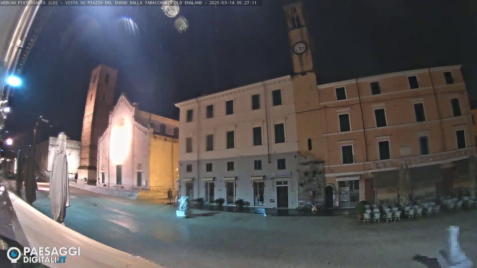 A nighttime view of a town square, showing a church with a tall bell tower, multi-story buildings, and a statue, with wet pavement suggesting recent rain or moisture.