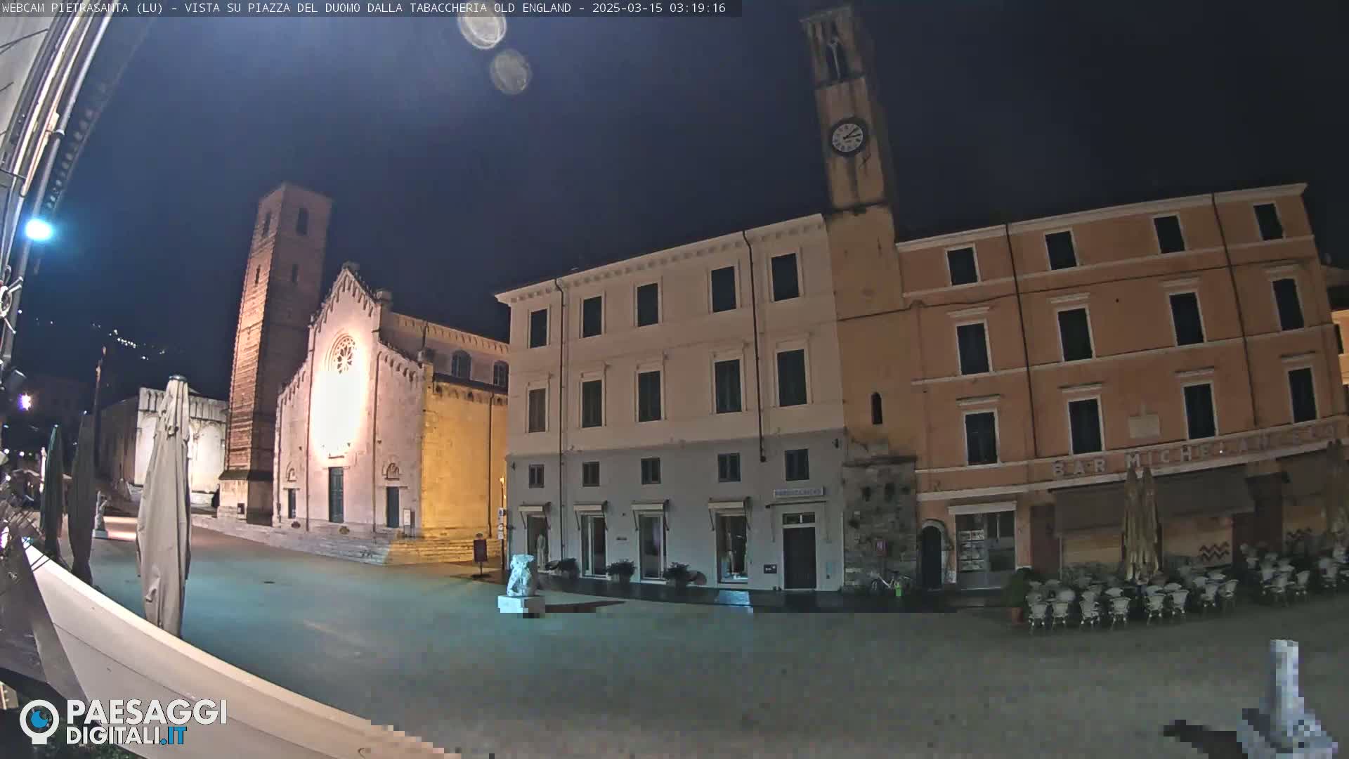 A nighttime view of a town square with several buildings, including a church with a tall tower and a clock tower, under a clear, dark sky.