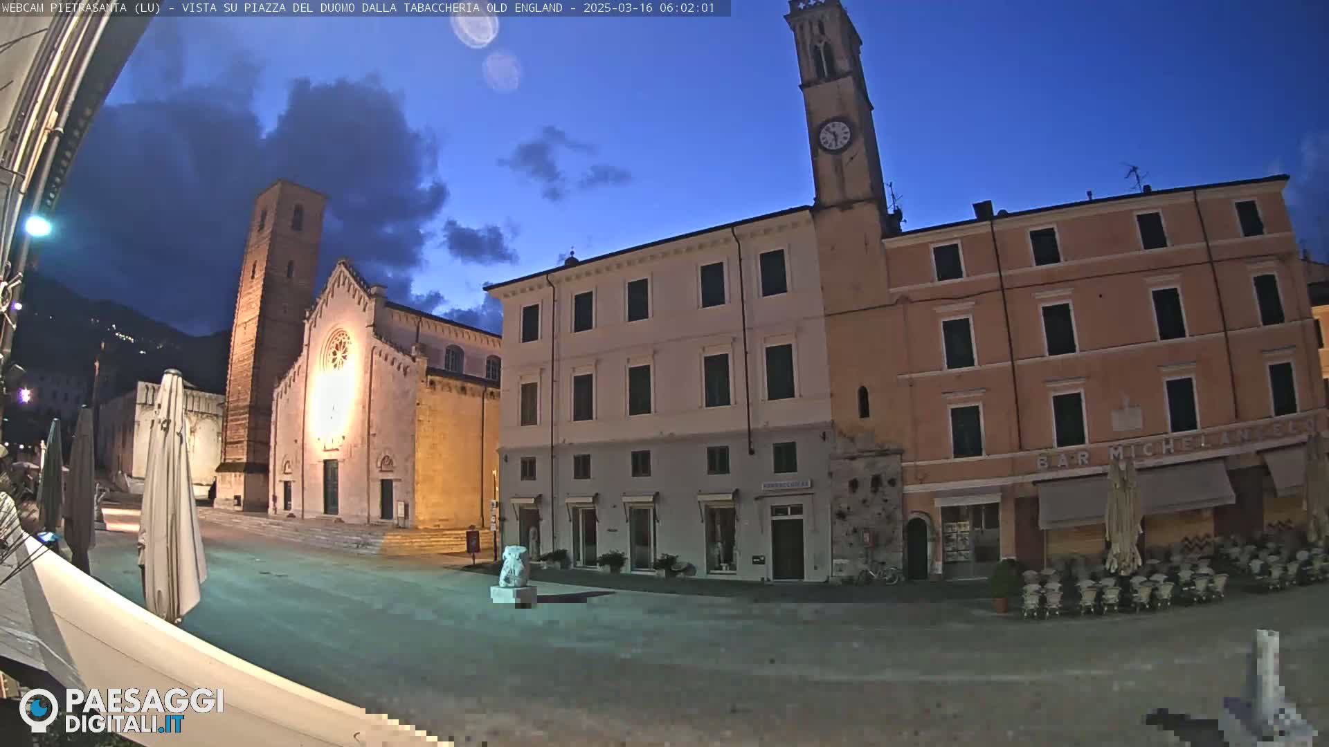 A town square at twilight, illuminated by artificial light, features a church with a tall bell tower, multi-story buildings, and outdoor seating areas.