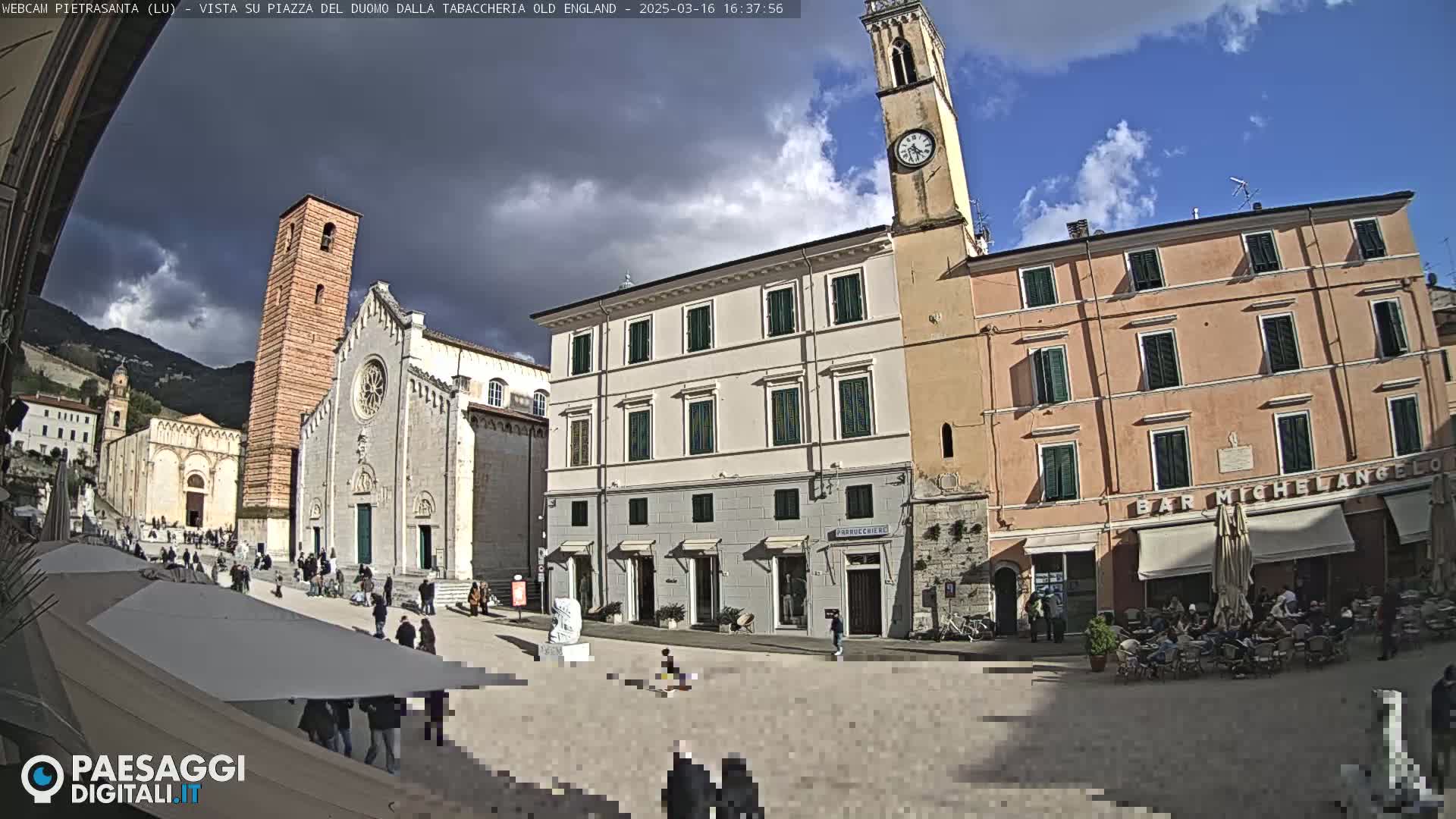 A partly cloudy sky hangs over a town square in Italy, where several historic buildings surround a plaza with people and outdoor seating.