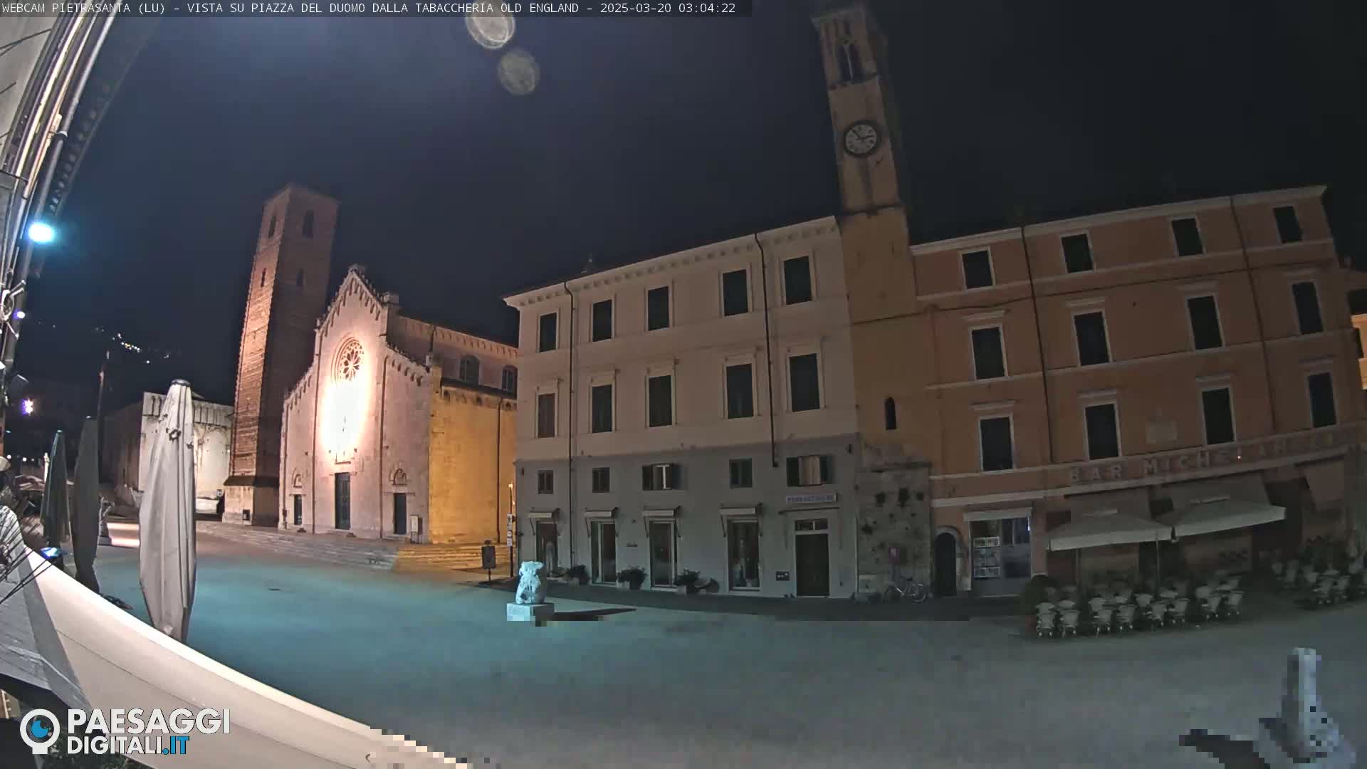 A nighttime view of a town square, featuring a church with a tall bell tower, several buildings, and a statue, under a clear, dark sky.