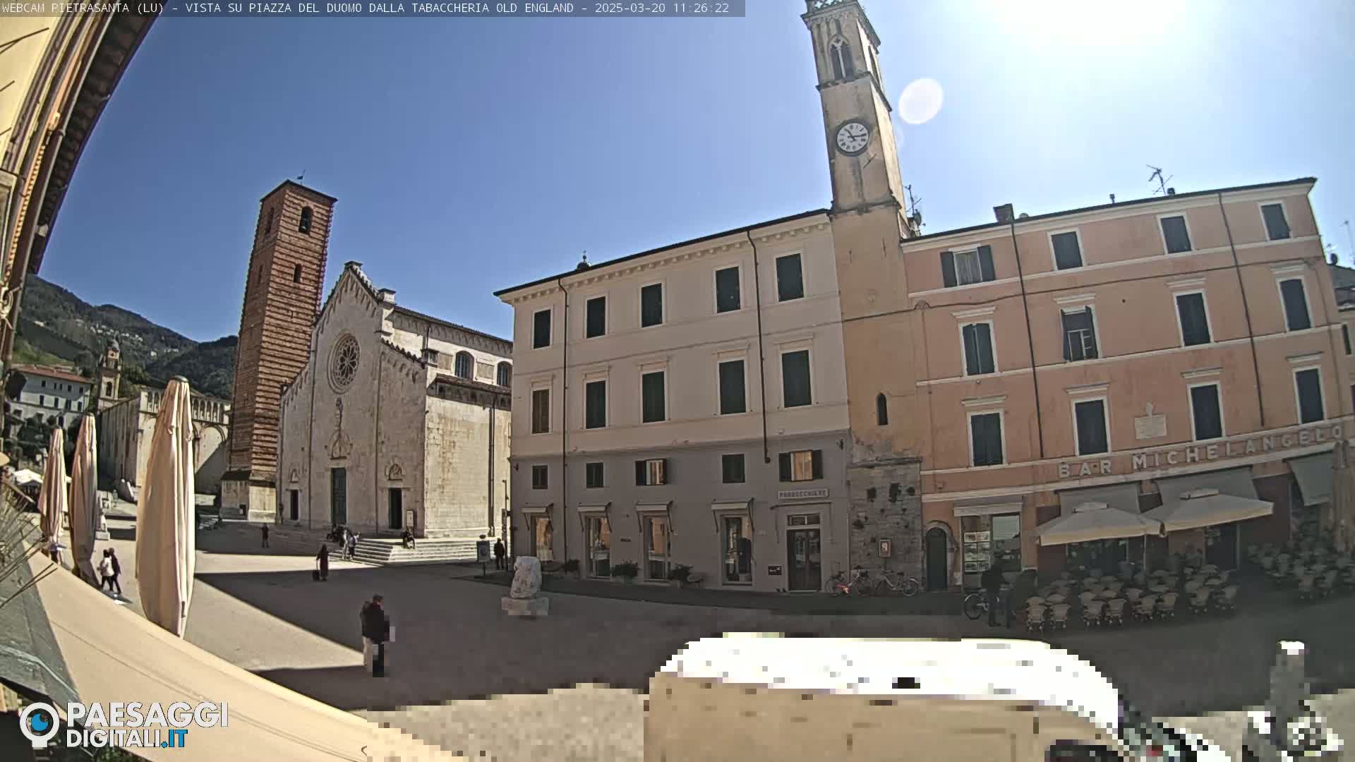 A sunny town square with a large cathedral, several buildings, and outdoor seating at a cafe.