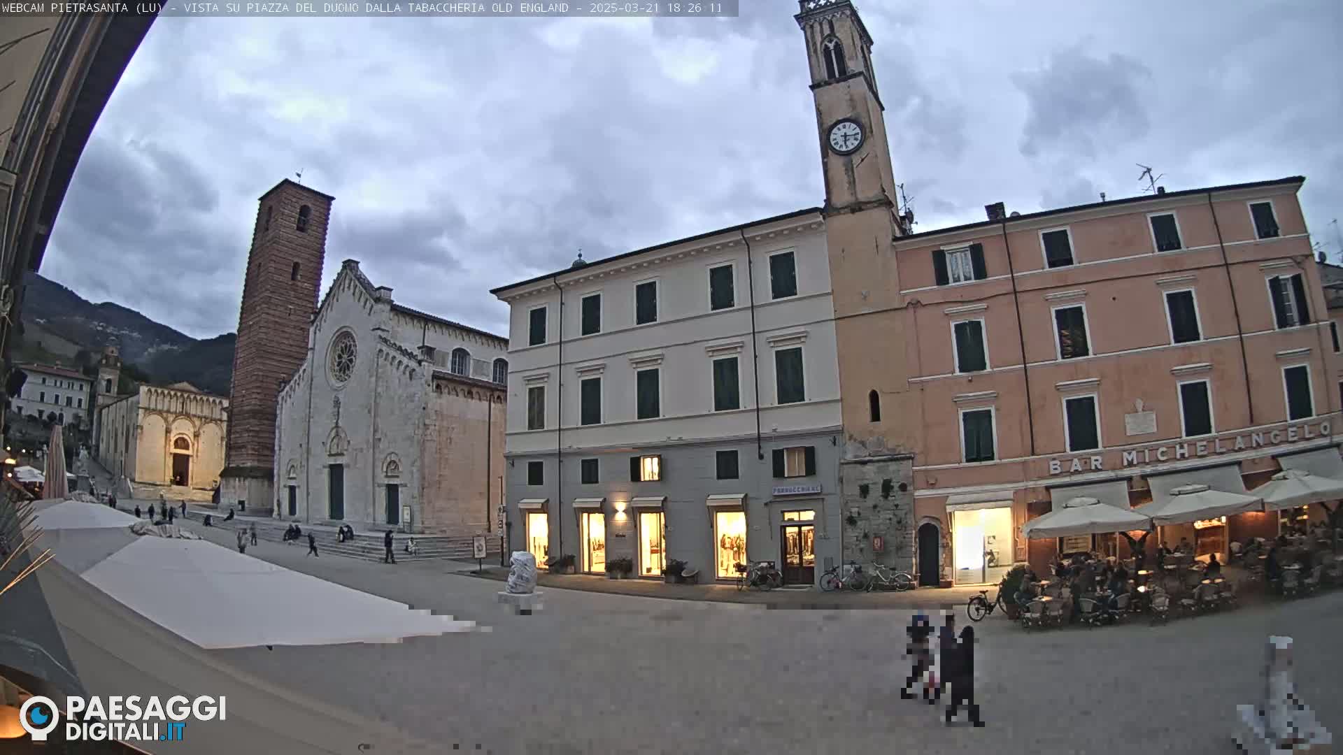A town square at dusk, under a cloudy sky, features a large cathedral, shops, and a clock tower, with a few people walking in the square.