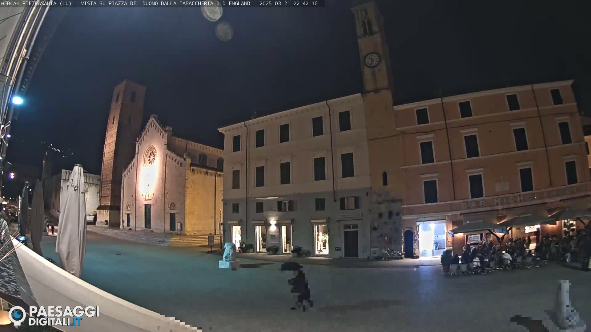 A nighttime view of a town square, illuminated by artificial light, featuring a church, buildings, and people sitting at outdoor café tables under umbrellas.