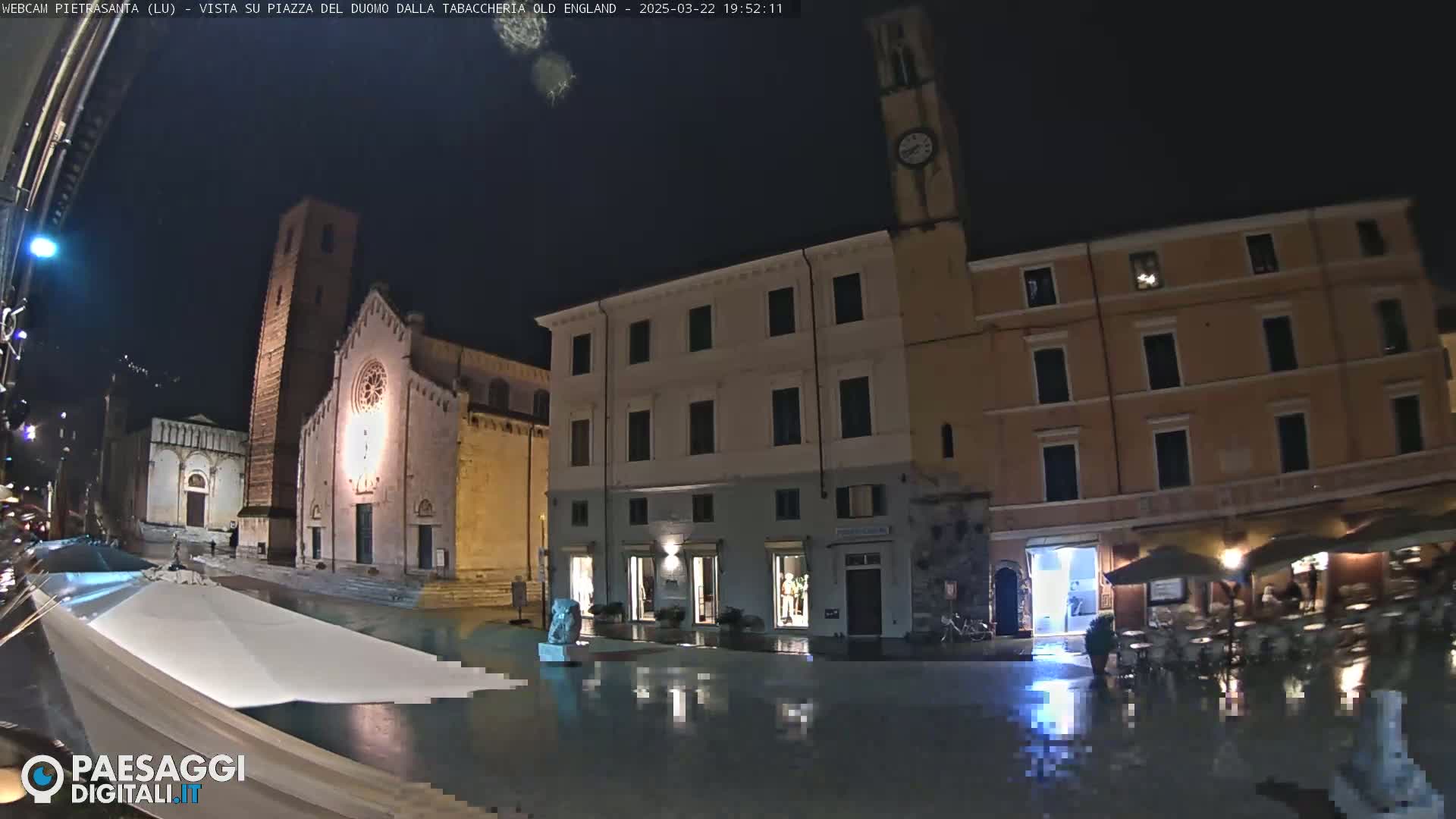 A rain-slicked town square at night is illuminated, showing a church, a clock tower, and buildings with outdoor seating areas.