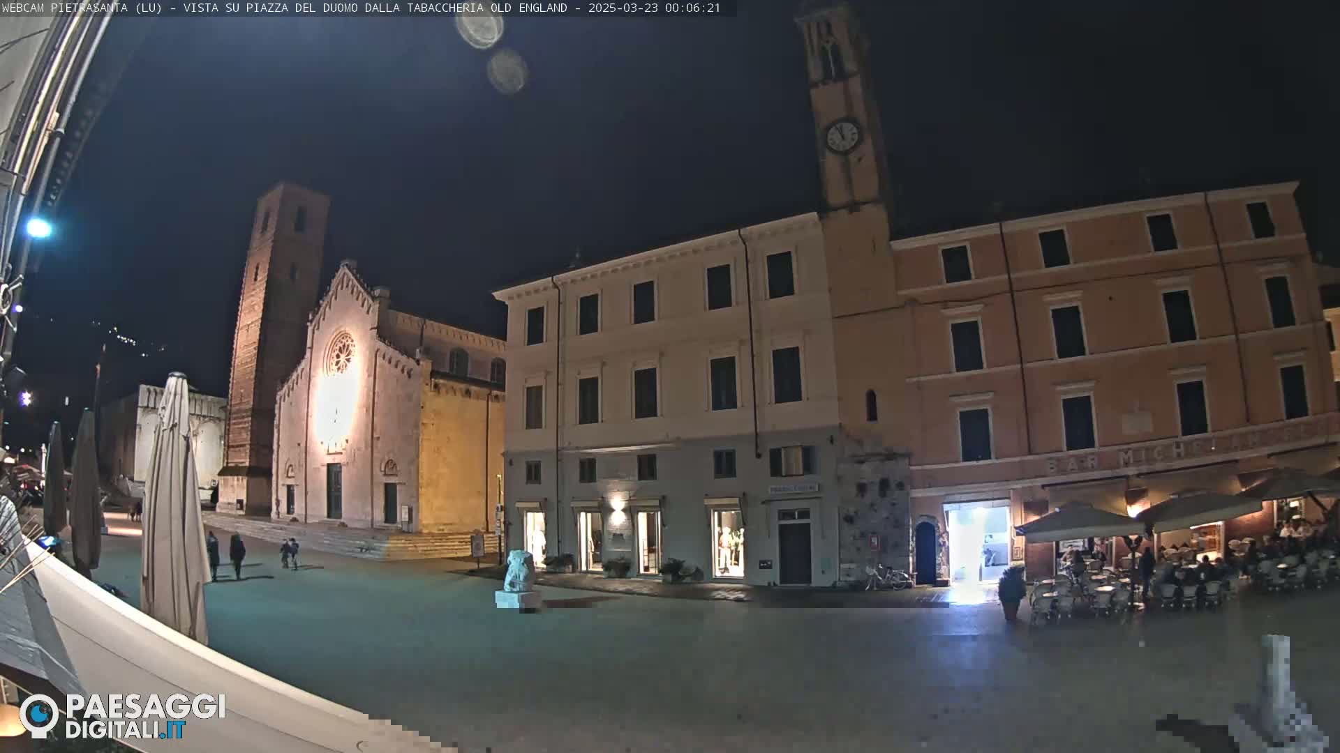 A nighttime view of a town square with a church, several buildings, and a few people; outdoor lighting illuminates the scene.