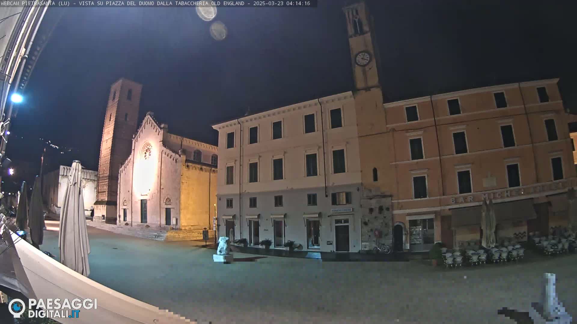 A nighttime view of a town square, featuring a church with a tall bell tower, several buildings, and a statue, under a clear, dark sky.