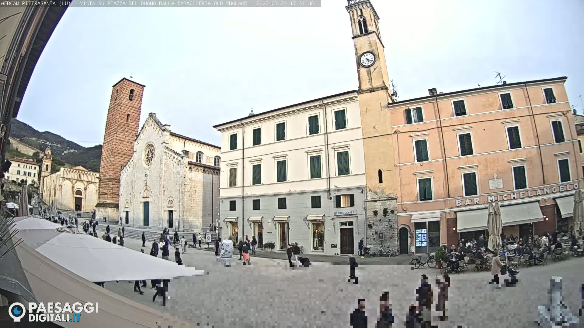 A town square, under a mostly clear sky, features several buildings including a cathedral and clock tower, with people walking around and sitting at outdoor cafes.