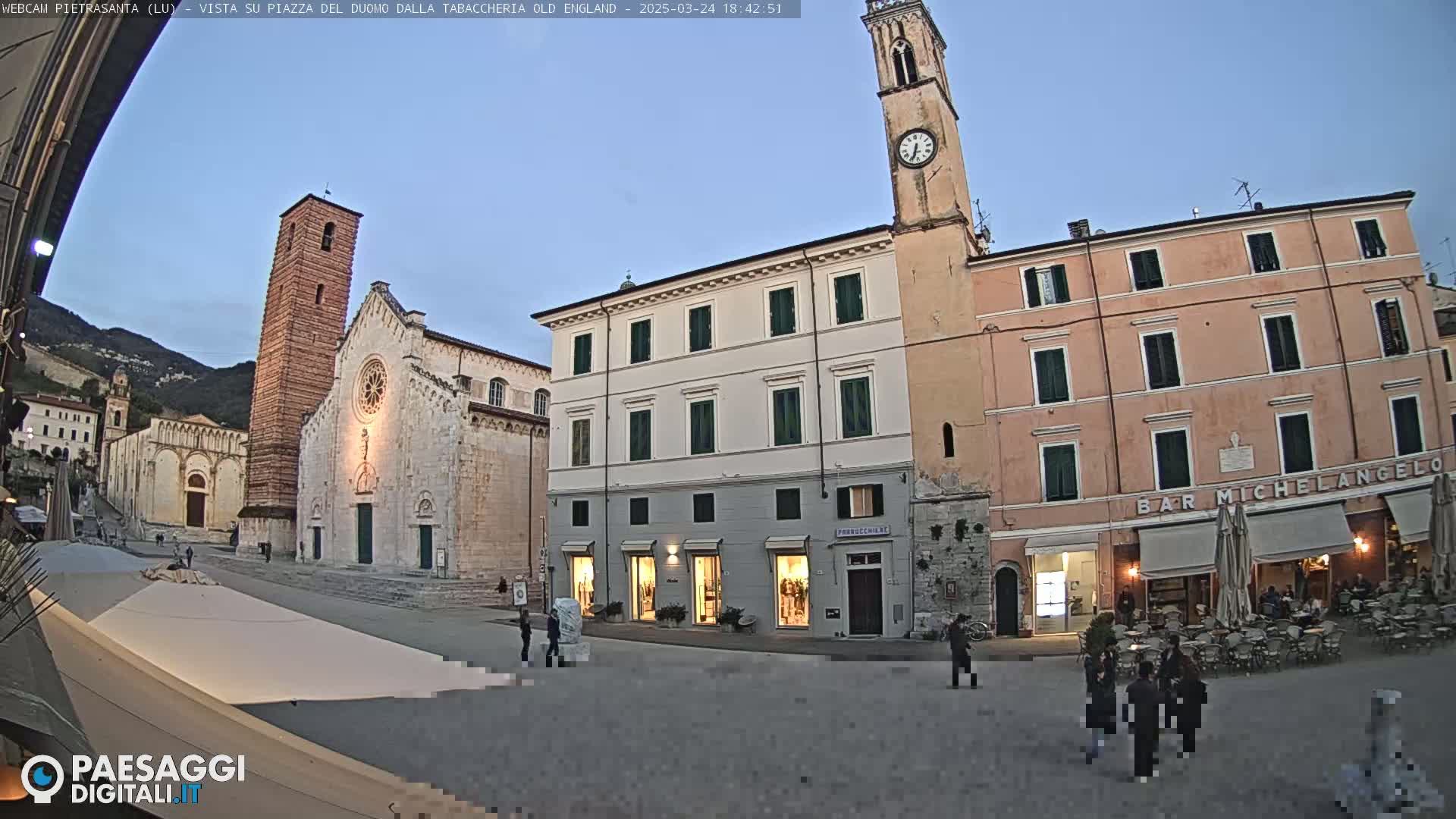 A town square at dusk, featuring a large church with a tall bell tower, adjacent buildings, and a few people strolling about.