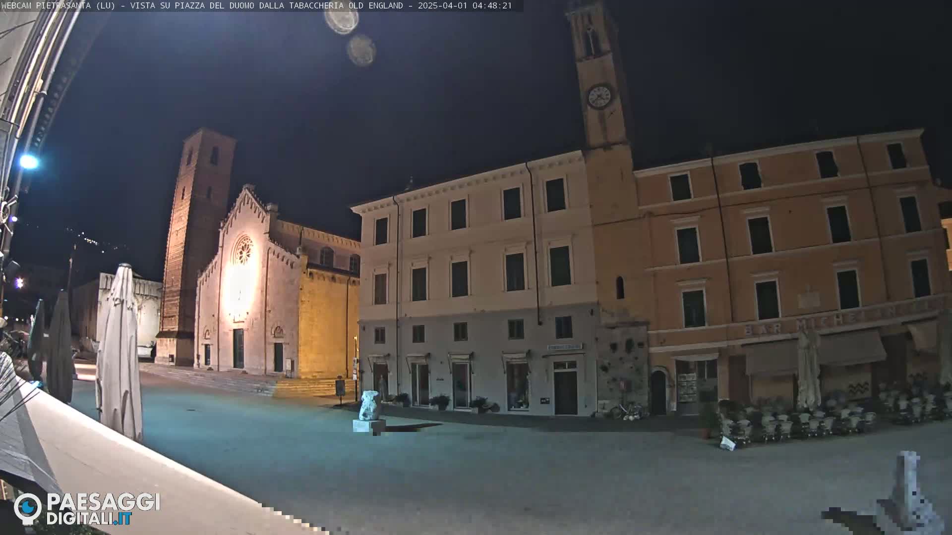 A nighttime view of a town square with a church, buildings, and a statue, under a clear, dark sky.