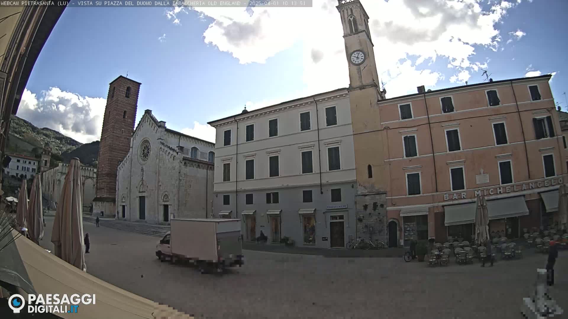A partly cloudy day in a town square shows a church with a tall bell tower, several buildings, and a few people; a delivery truck is parked in the square.