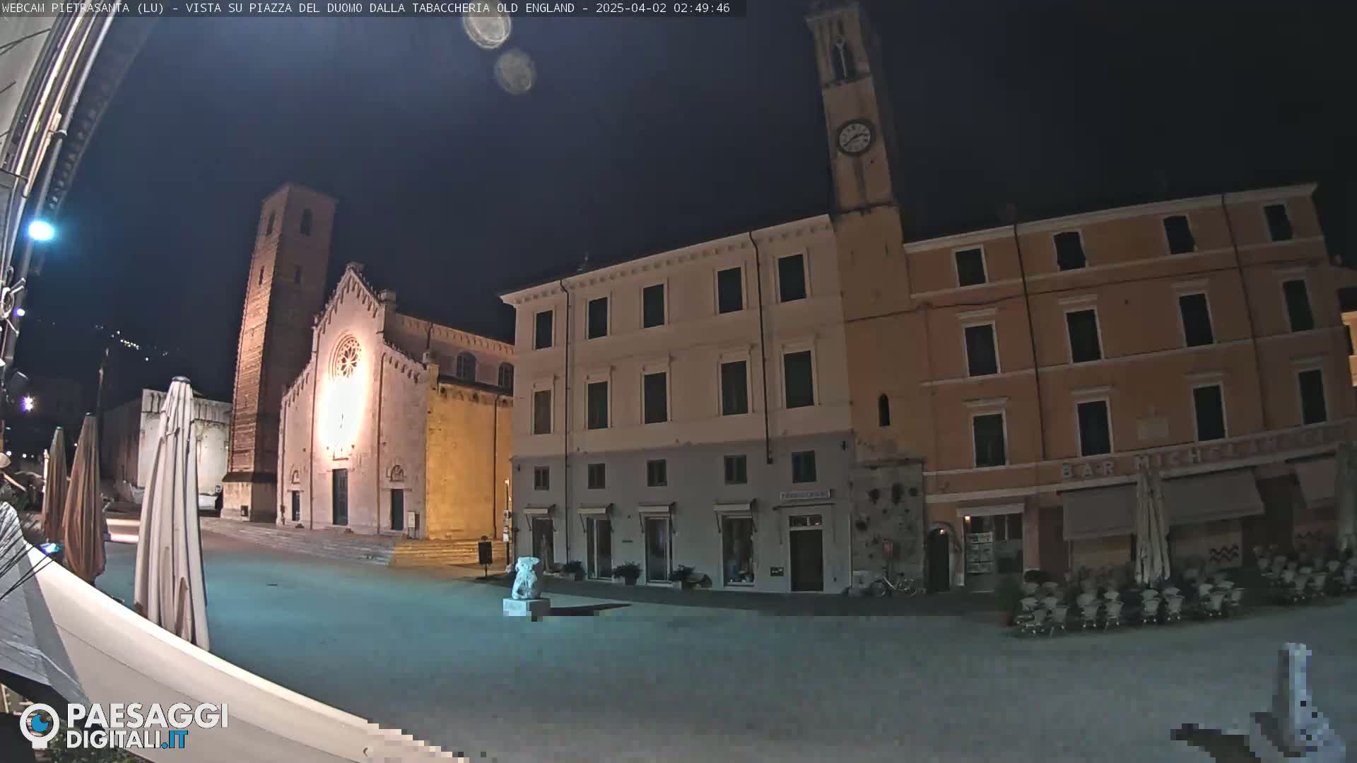 A nighttime view of a town square with a church, several buildings, and outdoor seating.