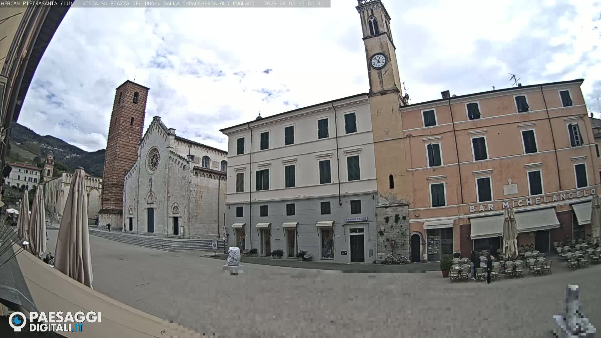 A town square with several buildings, including a church with a tall bell tower, under a partly cloudy sky.