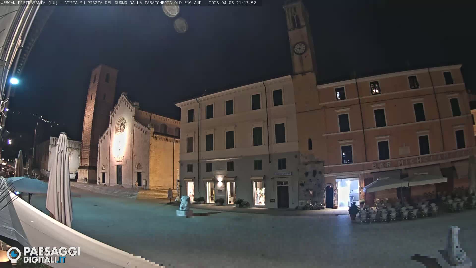 A nighttime view of a town square with a church, buildings, and outdoor seating.