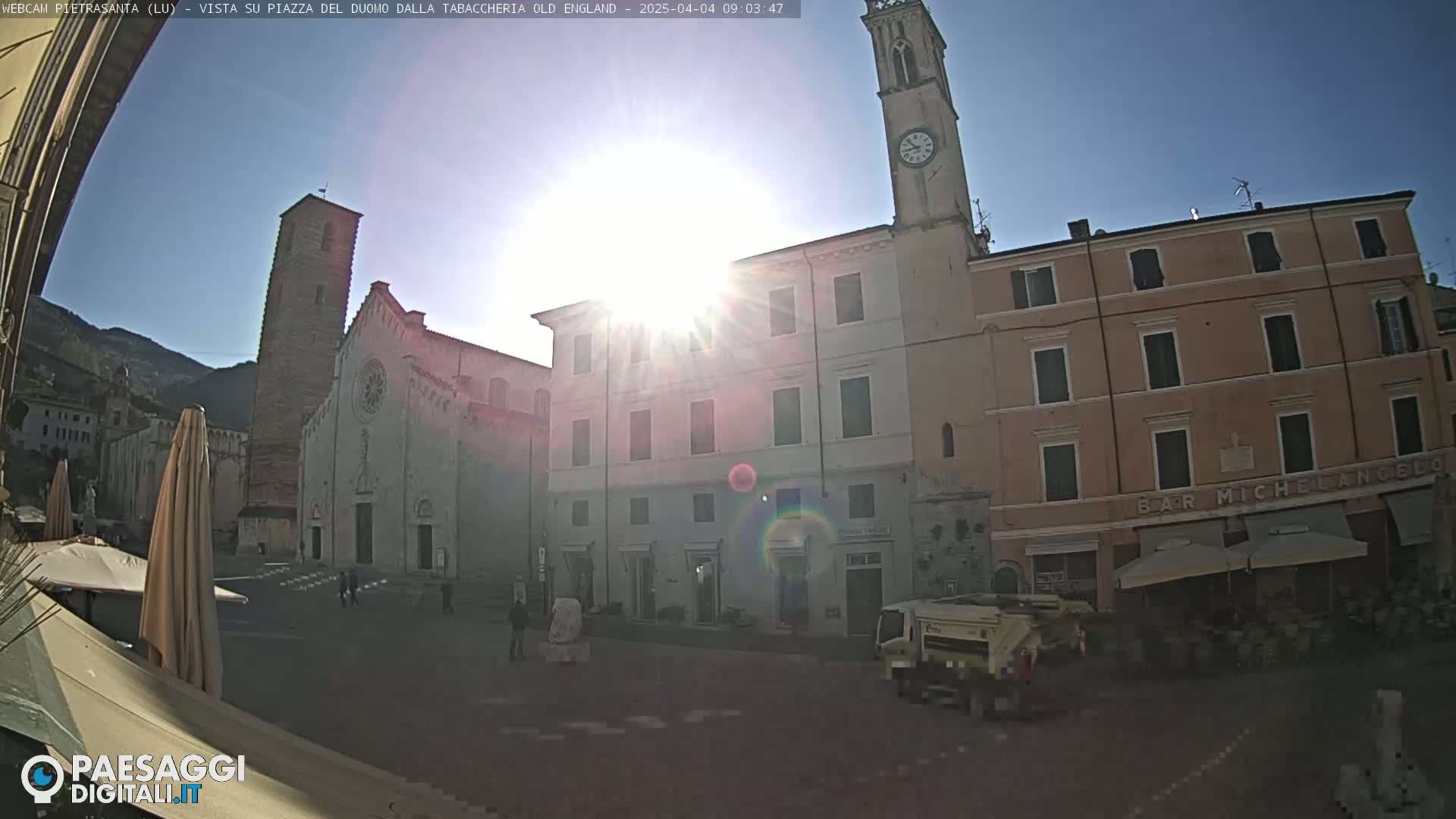 A sunny town square is shown, featuring a large church with a tall bell tower, several multi-story buildings, and a few people walking.