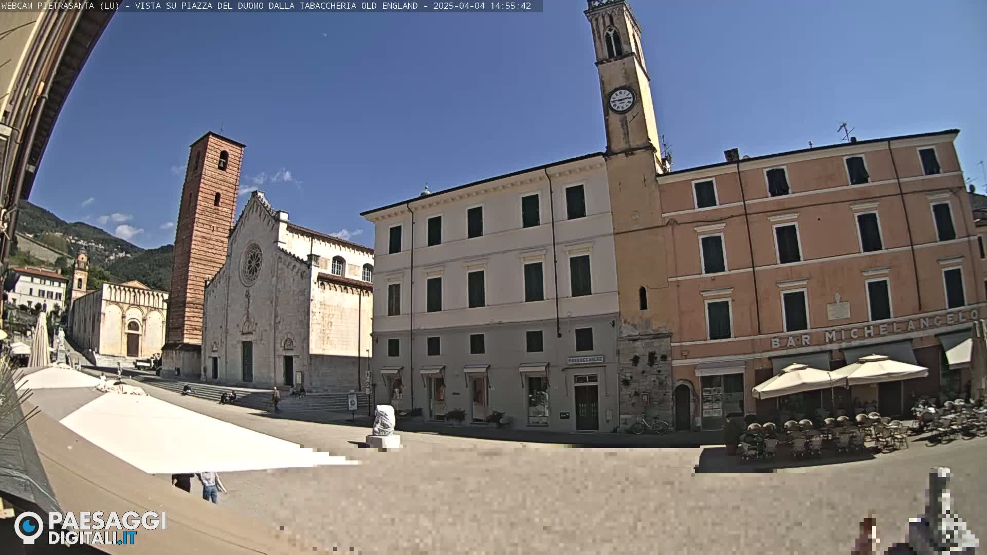 A sunny town square with several buildings, including a church with a tall bell tower, and a café with outdoor seating.