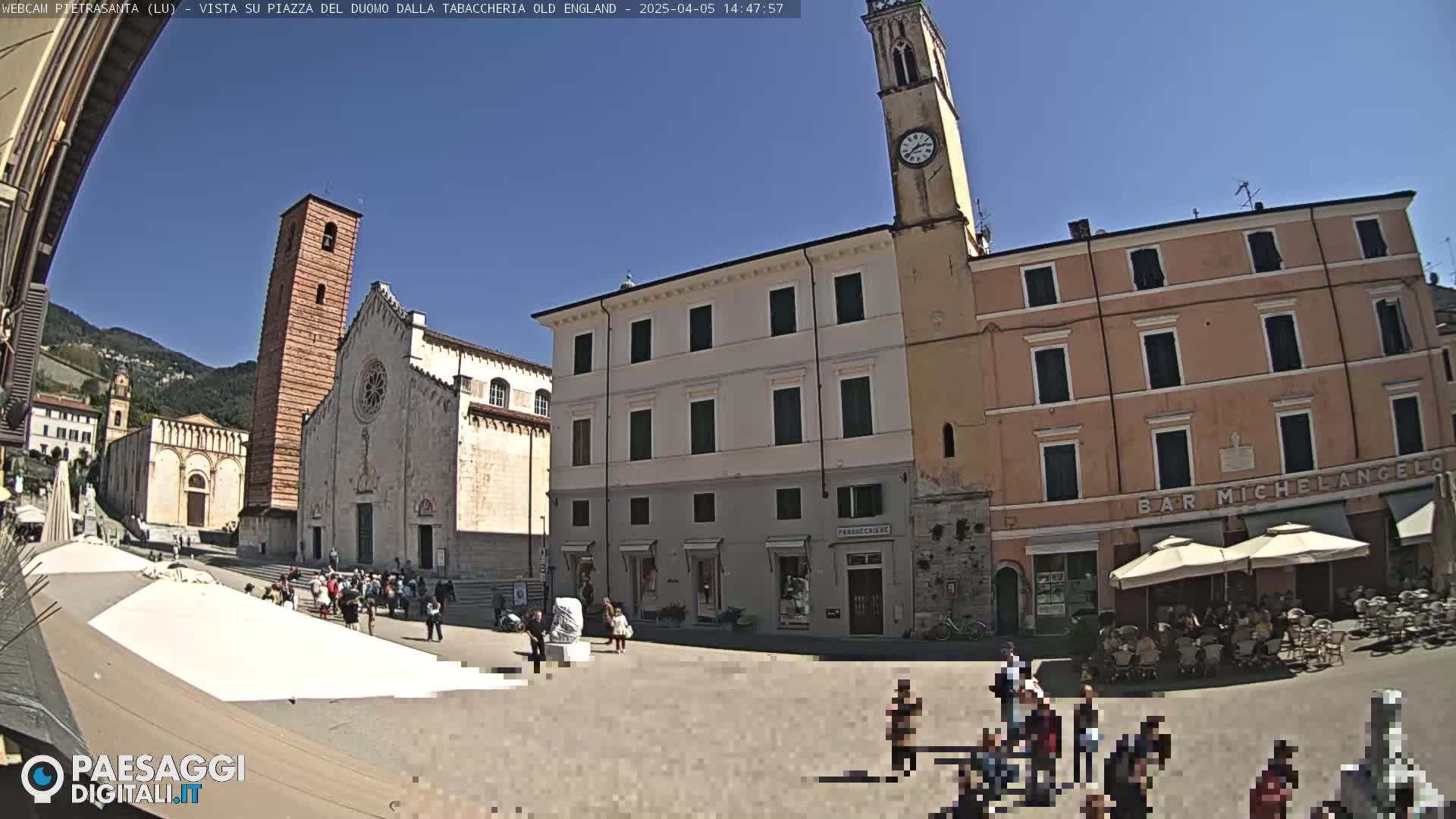 A town square in bright sunlight, featuring a large church, several buildings, and people gathered in and around outdoor cafes.