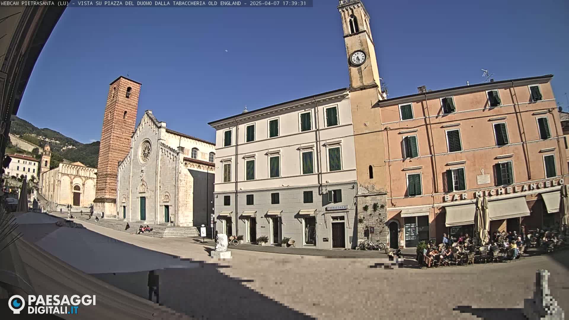 A sunny town square is shown, featuring a large cathedral, several buildings, a clock tower, and an outdoor cafe with patrons seated at tables.