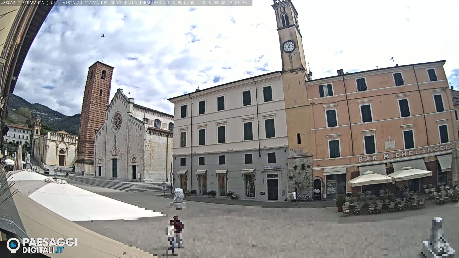 A partly cloudy day reveals a town square with a large cathedral and several buildings, including a cafe with outdoor seating.