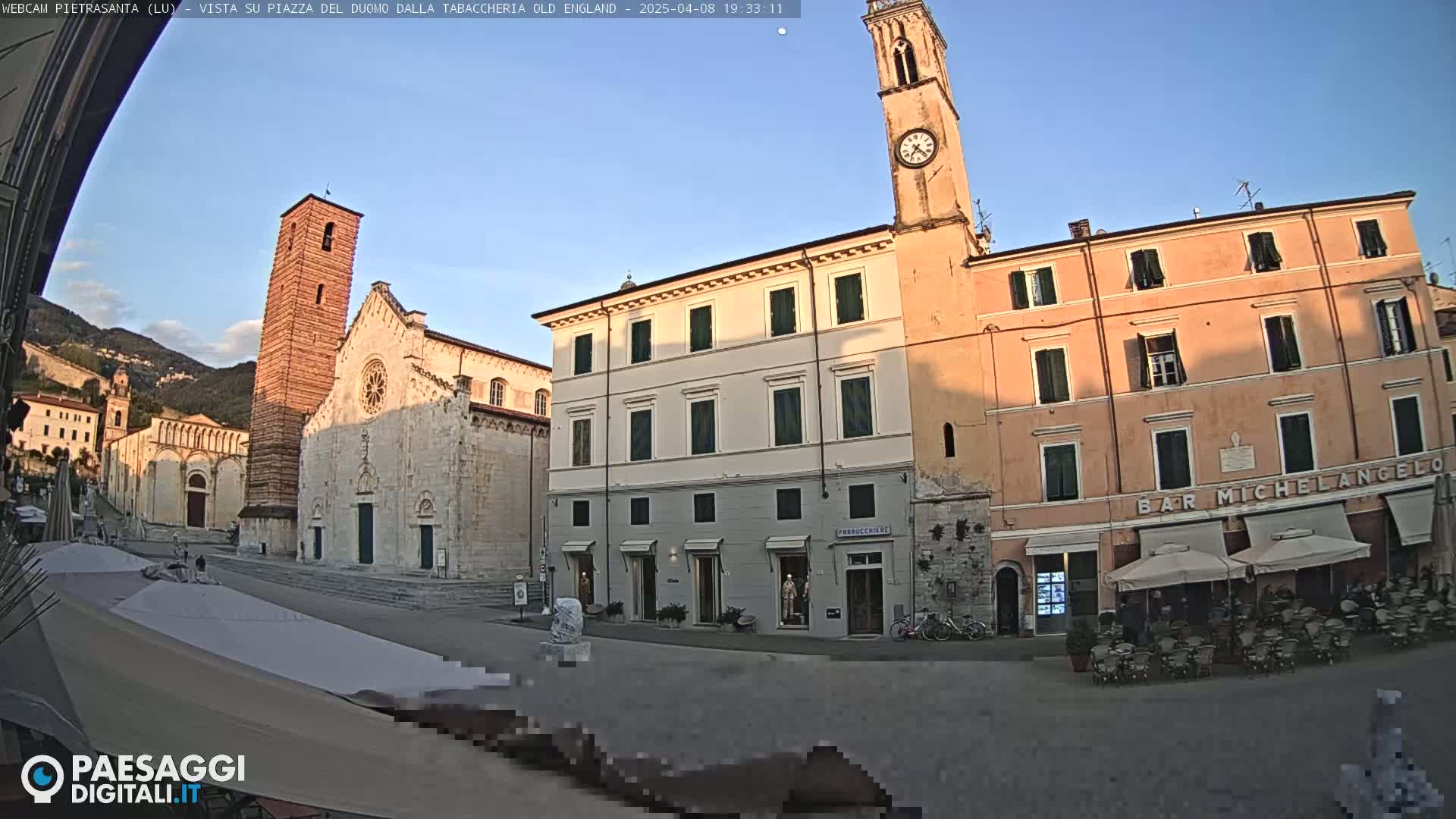A sunlit town square features a large stone church with a tall bell tower, adjacent buildings, outdoor cafes with umbrellas, and a background of distant mountains under a clear sky.