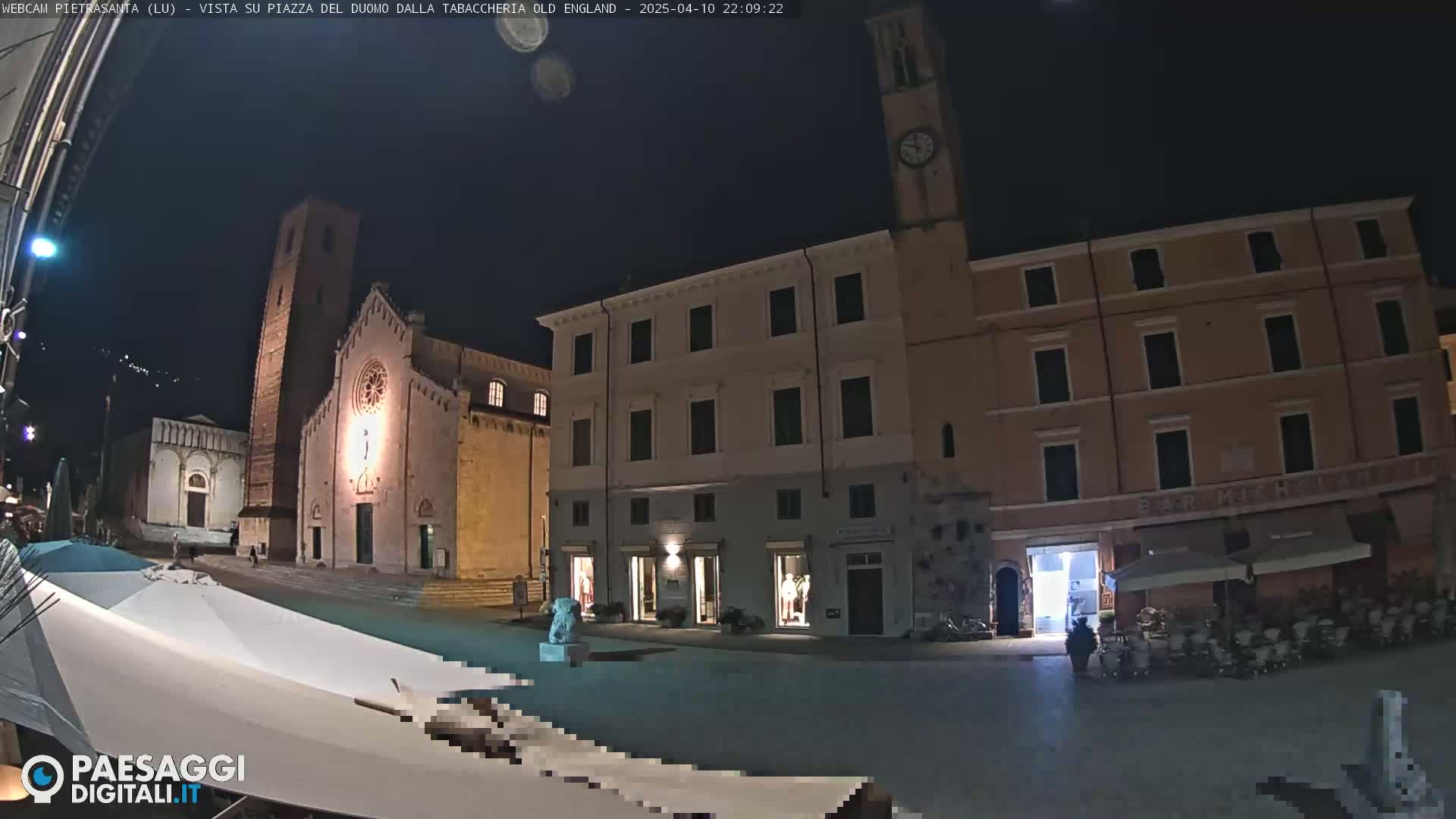 A nighttime view of a town square with a church, buildings, and outdoor seating at cafes under a clear sky.