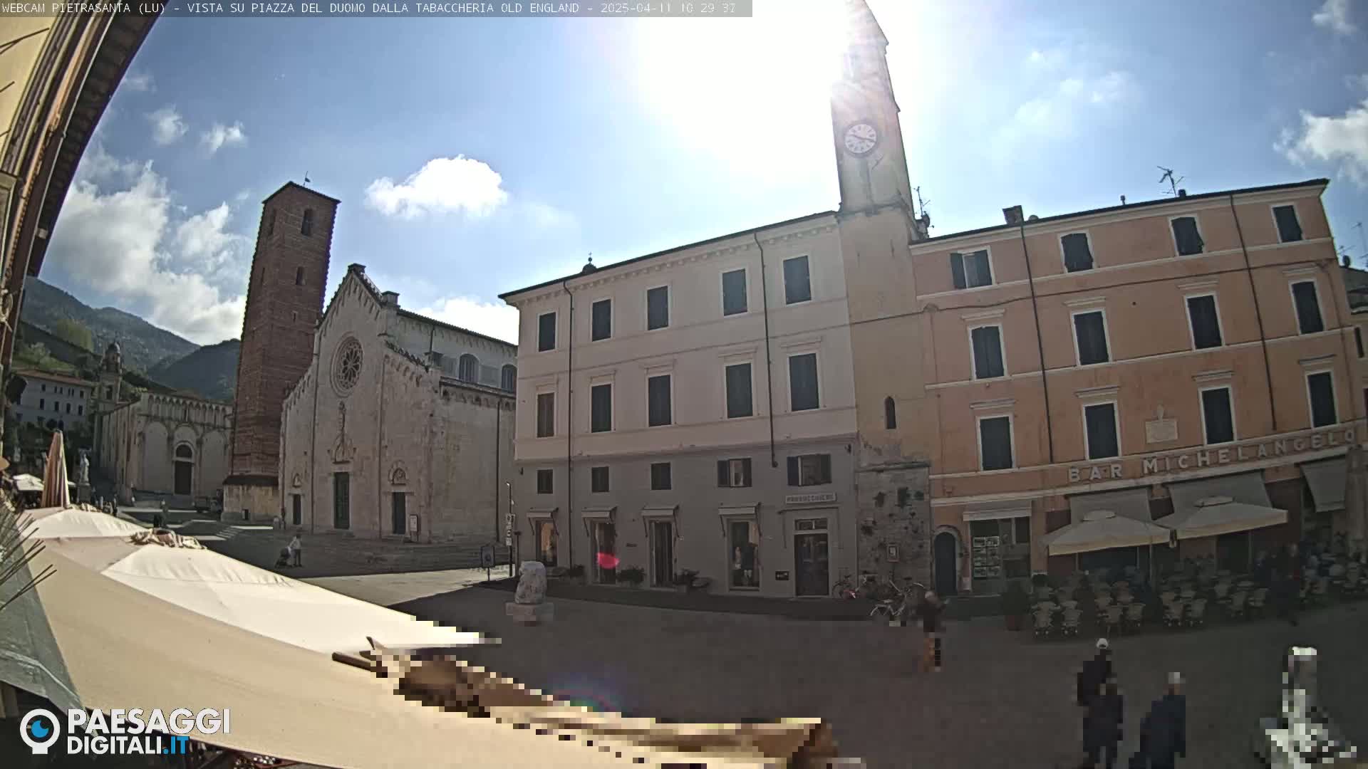 A sunny town square is shown, featuring a large cathedral, multi-story buildings, and a few people milling about.