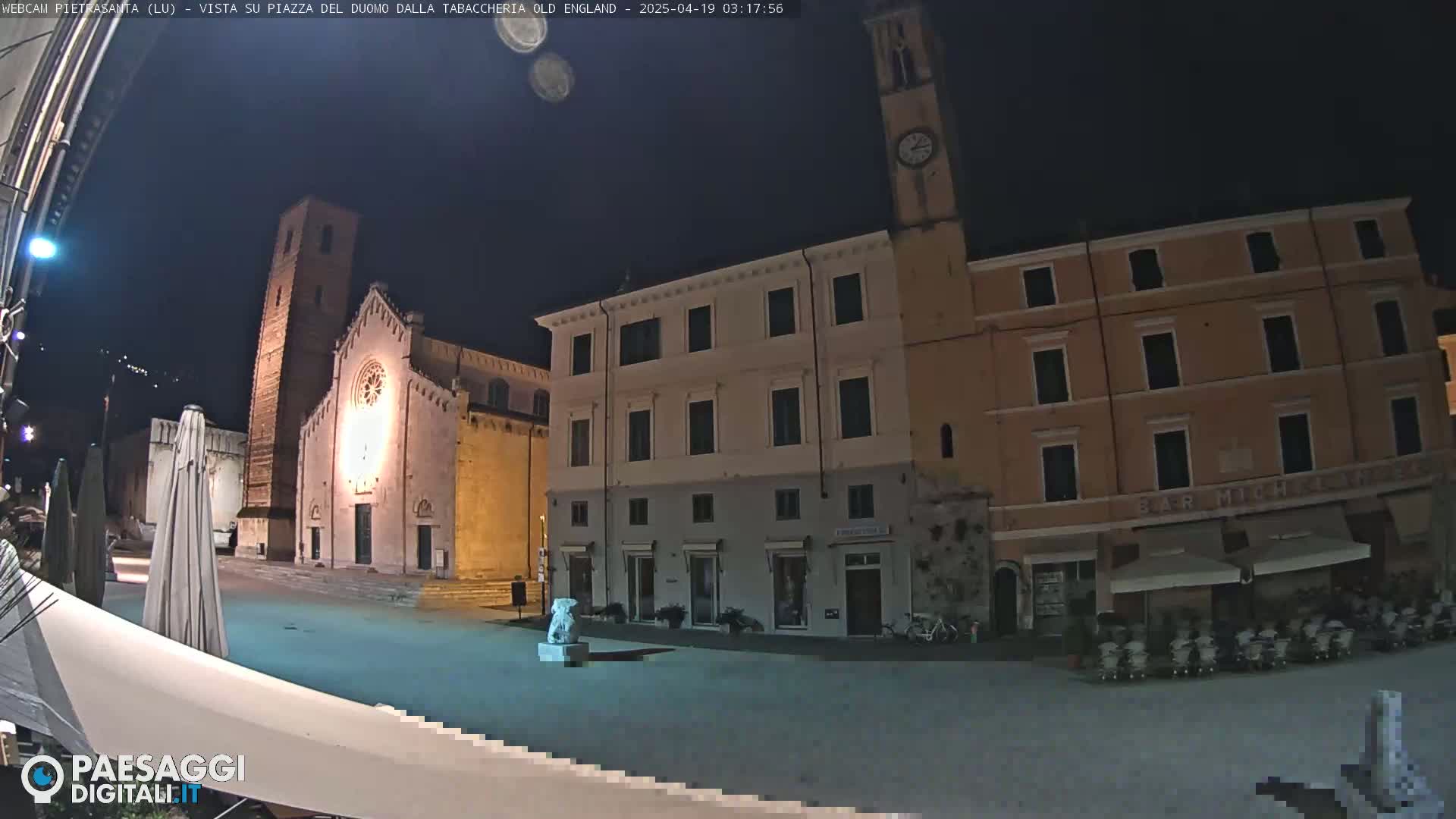 A nighttime view of a town square, featuring a church, several buildings, and a statue, under a dark sky.